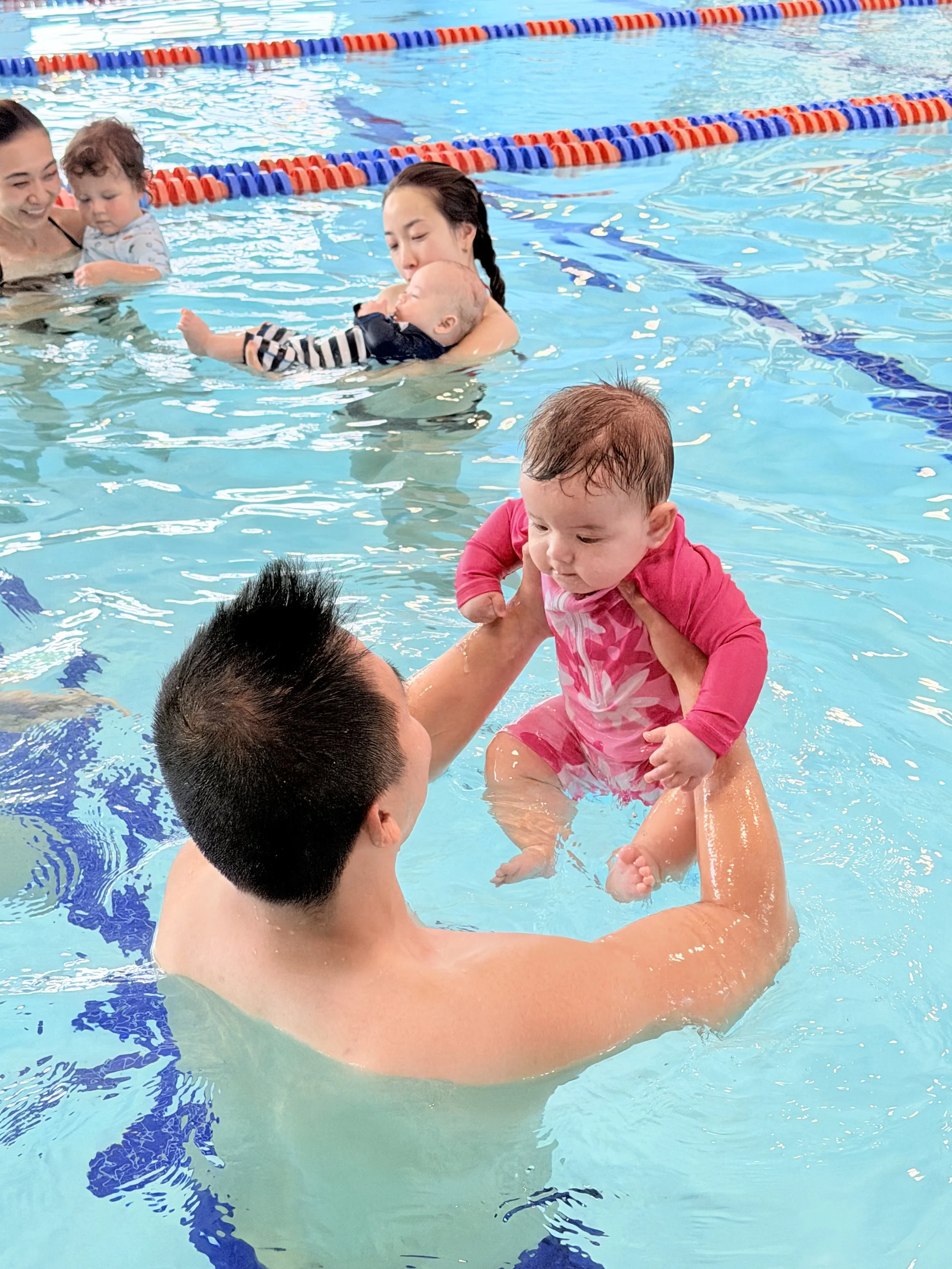 A swimming pool scene with young children and adults, some are holding children, and the pool has multicolored lane dividers.