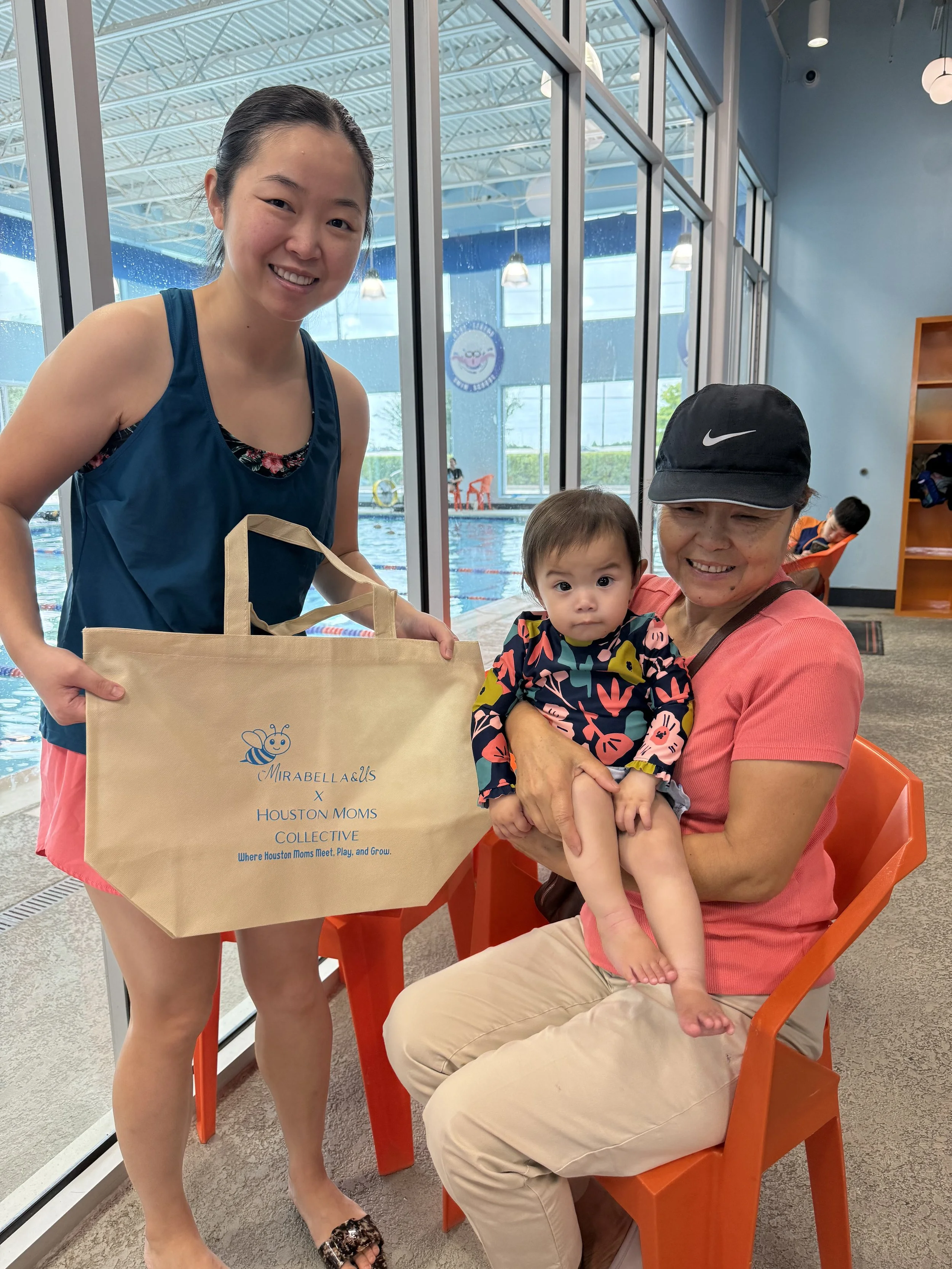 A woman holding a young child while sitting on an orange chair inside a swimming pool facility, a woman in a blue tank top presenting a beige tote bag, and another woman in a black Nike hat. There are large windows and a swimming pool in the backgrou