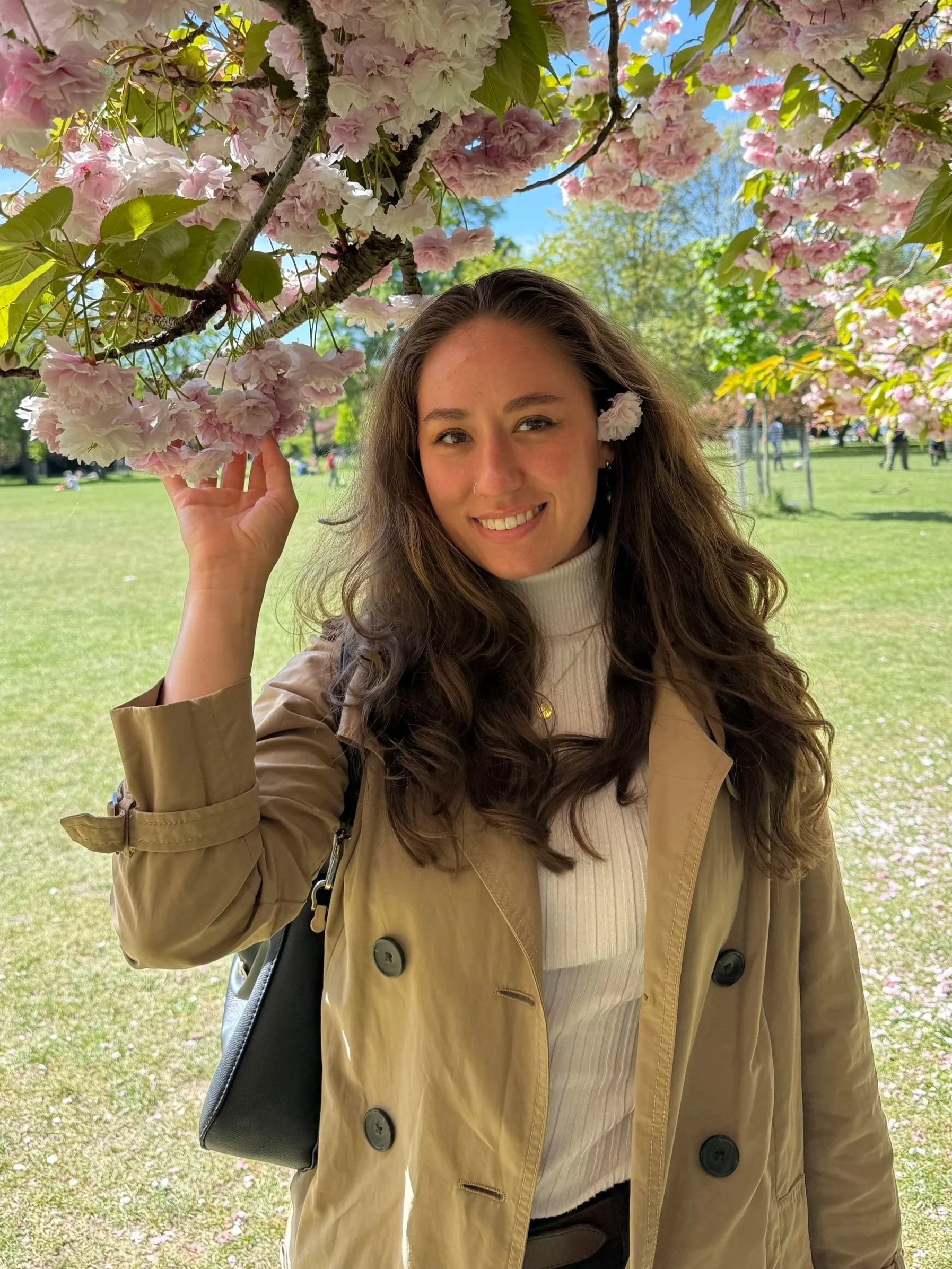 A young woman with long wavy hair smiling outdoors in a park, standing under a blossoming pink cherry blossom tree on a sunny day, wearing a beige trench coat and a white turtleneck.