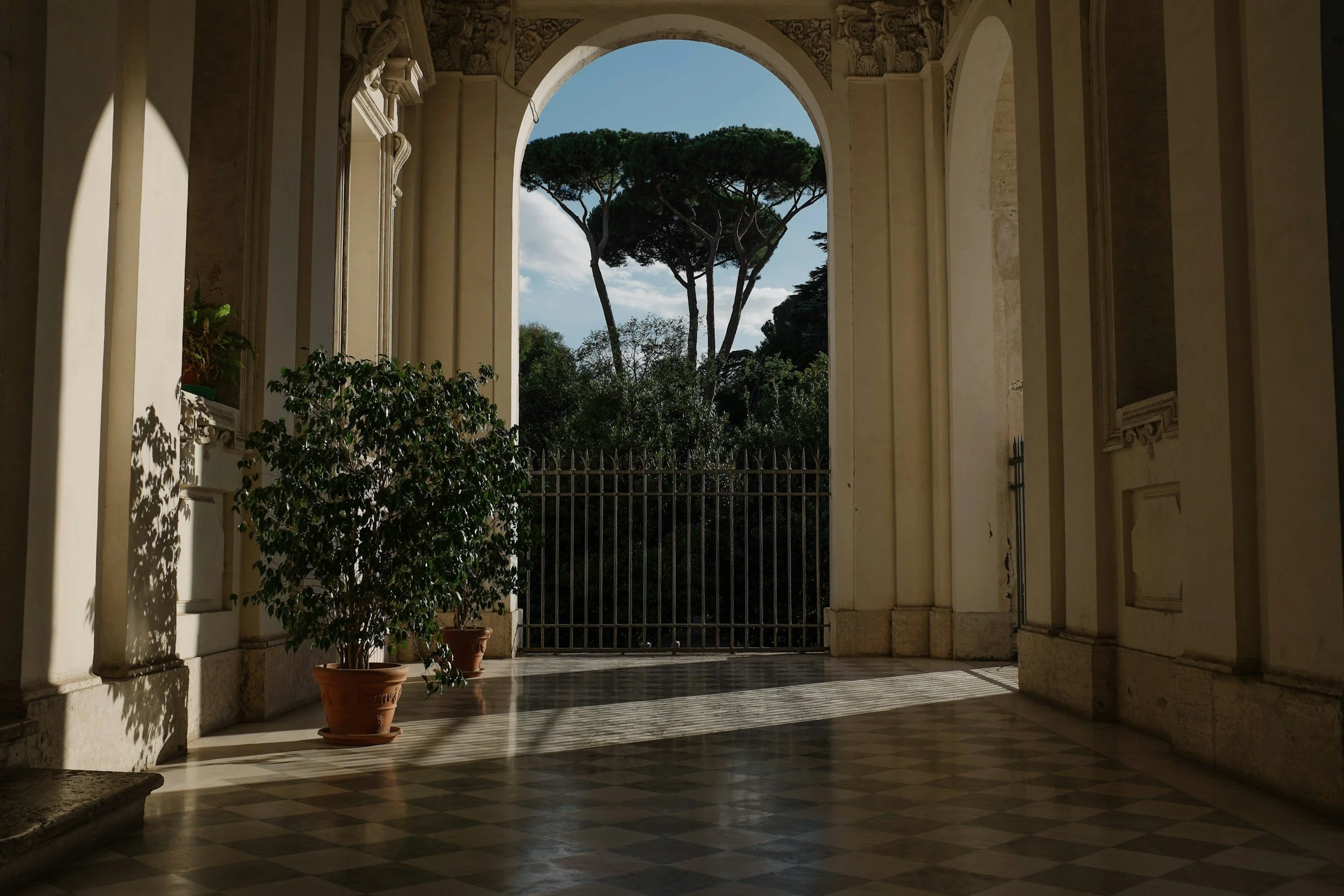 A view through a large arched window in a historic building shows trees and blue sky with clouds outside. The interior has decorative architectural details, potted plants, and a checkered marble floor.