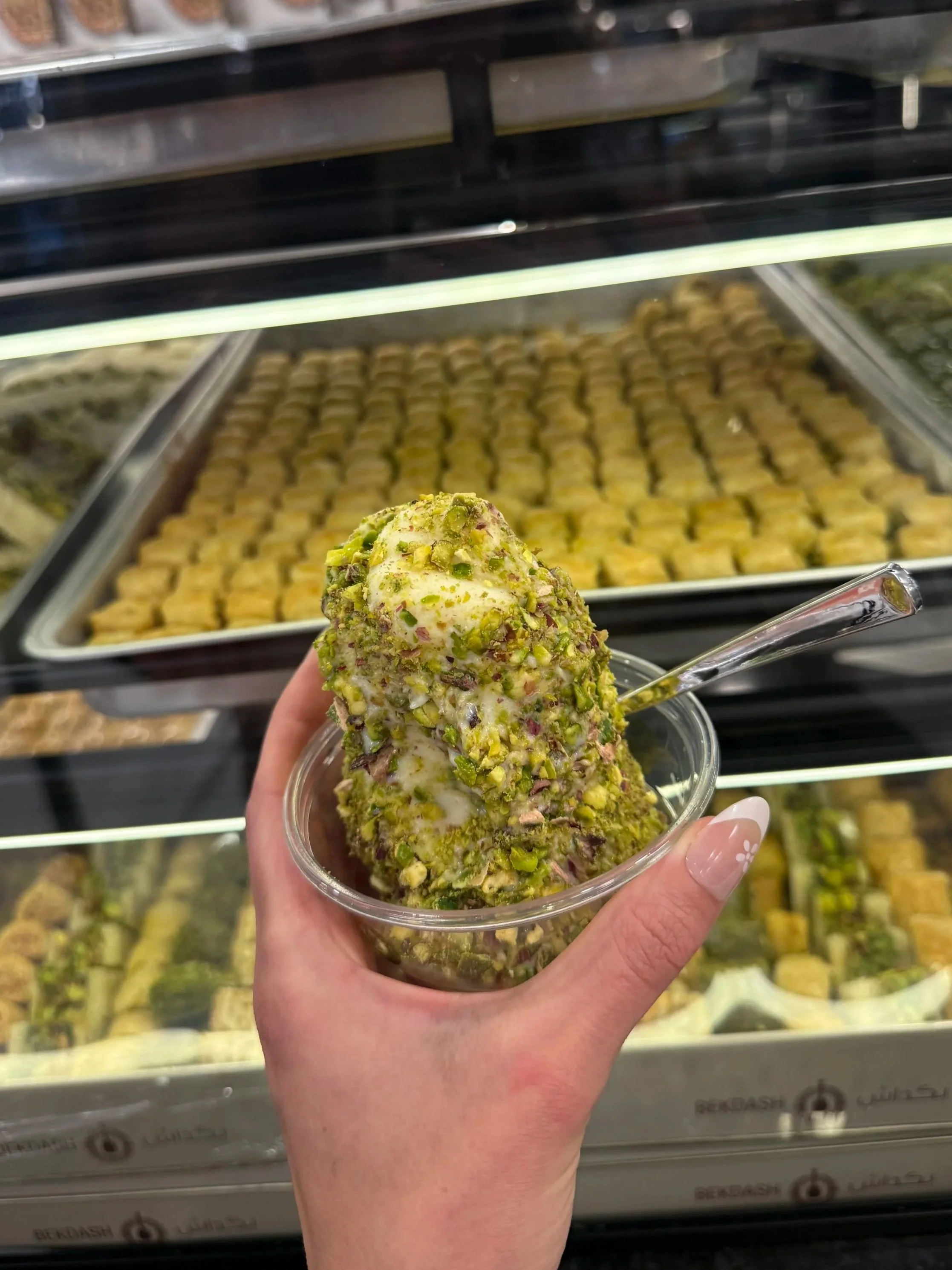 Hand holding a cup of pistachio-flavored ice cream with pistachio pieces on top, in front of a display case filled with various desserts.