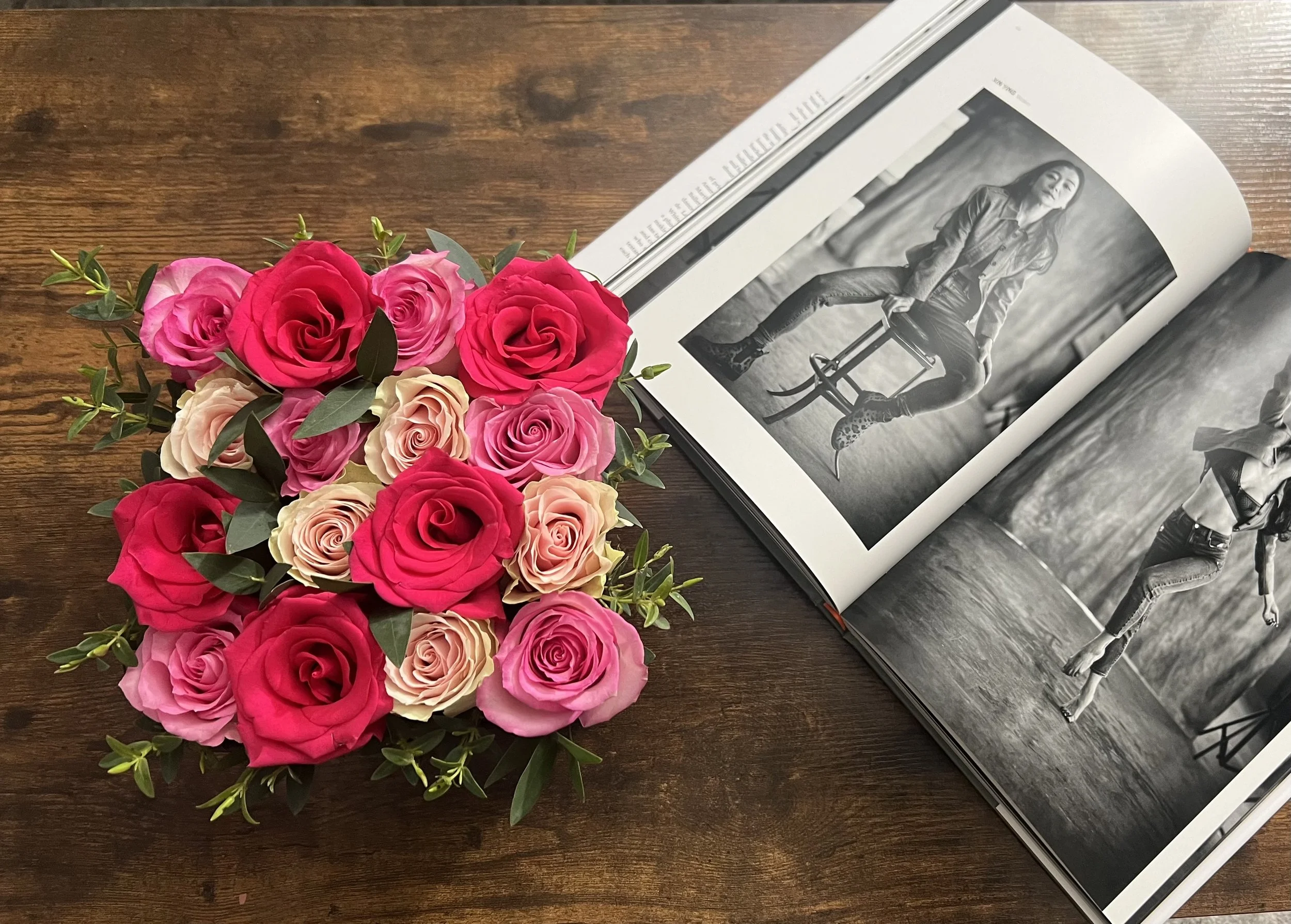 A bouquet of pink and cream roses with green leaves on a wooden table next to an open black and white photo book with photos of a woman posing in casual clothing.