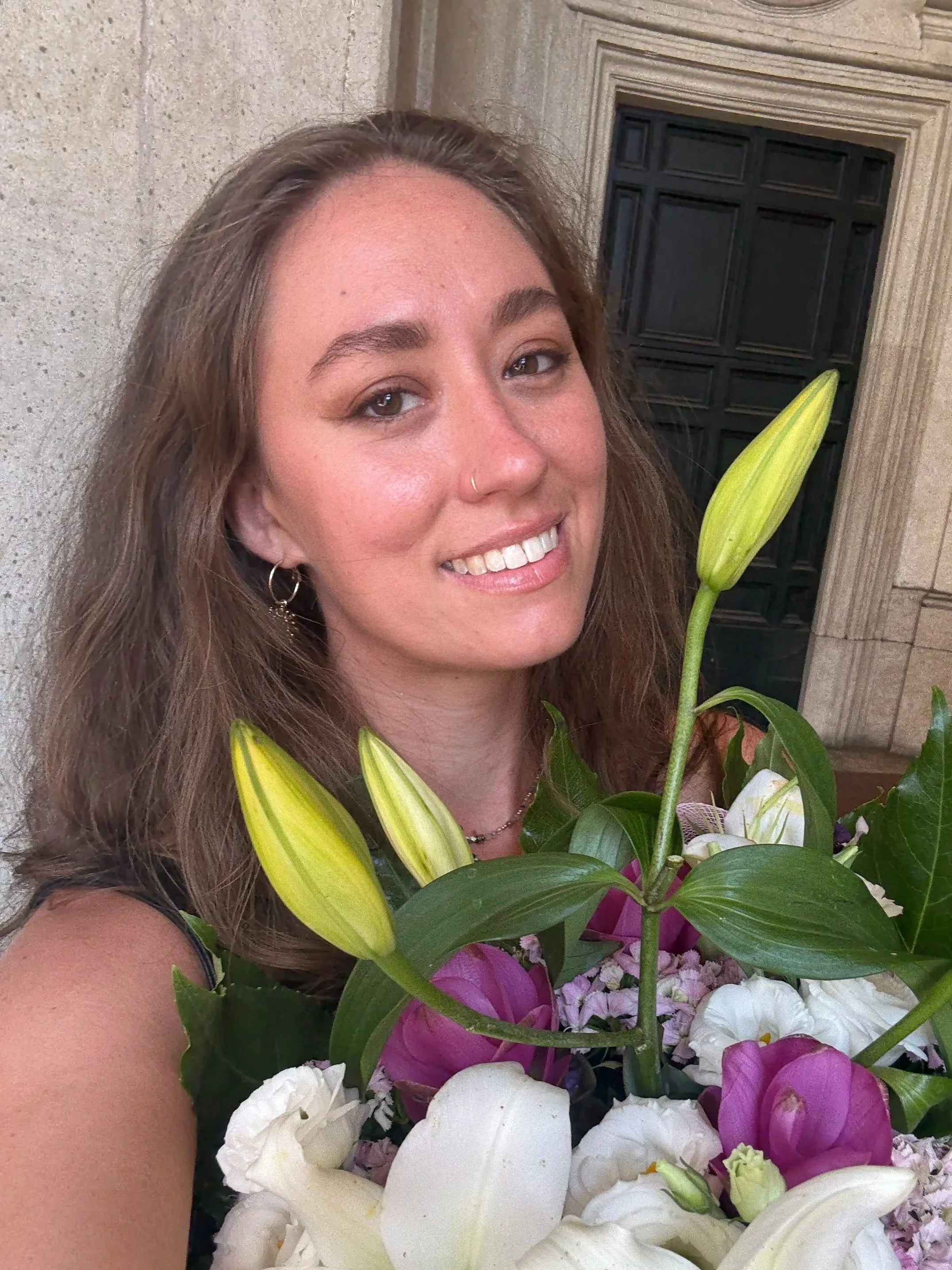 A woman with long brown hair, smiling, holding a bouquet of colorful flowers, including lilies, roses, and other blossoms, in front of a stone building with a black window.