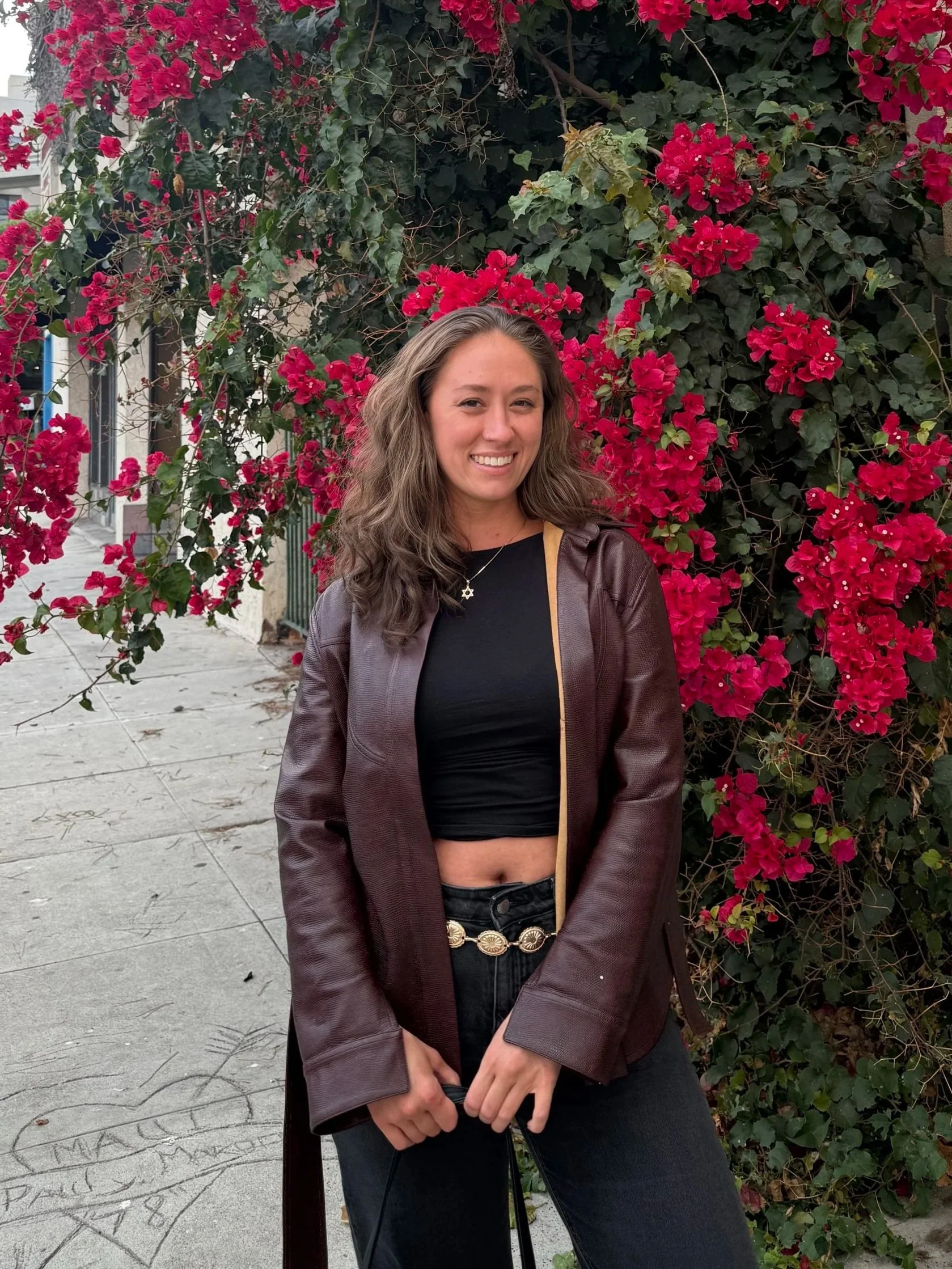 A young woman with wavy brown hair, wearing a black crop top, a brown leather jacket, and a necklace with a star pendant, standing in front of a large bush with bright pink flowers.