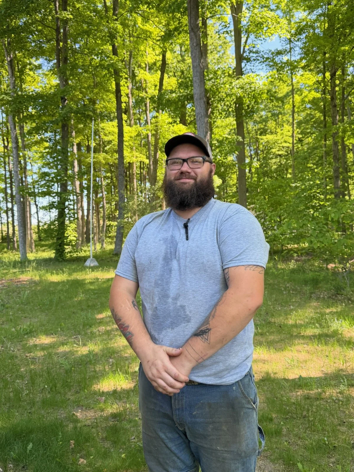A man with a beard wearing a gray hoodie and gray sweatpants, standing in front of a tree in a forest during daytime, smiling at the camera.