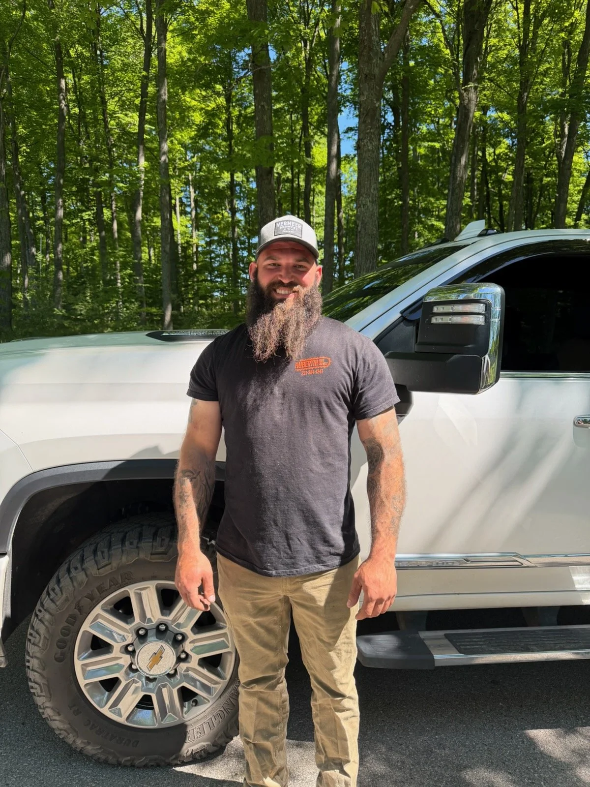 A man with a beard wearing a gray cap and black shirt with an outdoor company logo, standing next to a tree in a lush green forest.