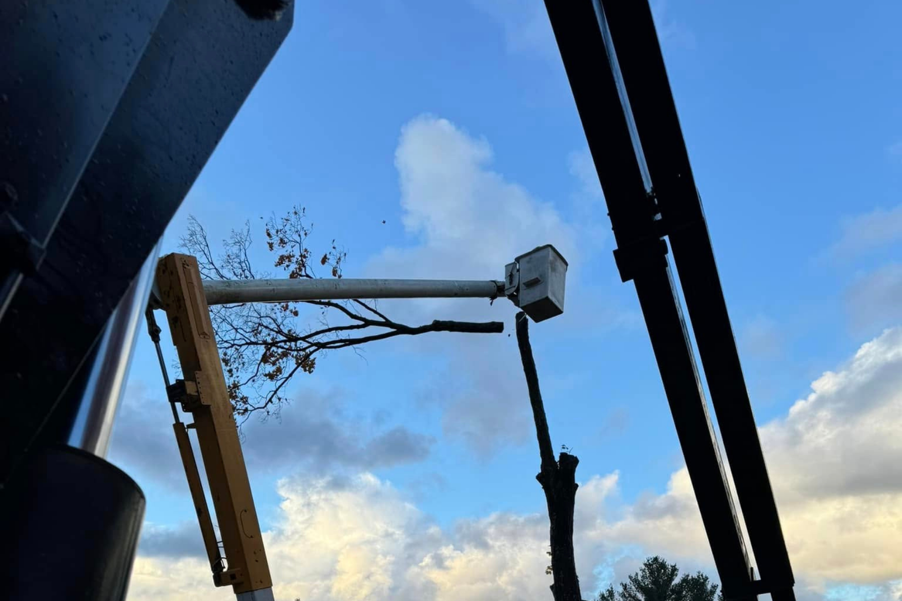 A utility bucket lift reaching toward a tree with some bare branches, with a blue sky and clouds in the background.