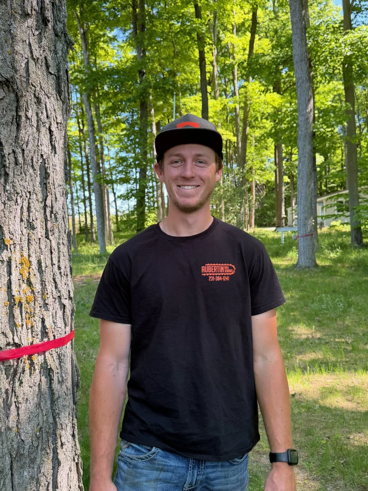A man with a full beard, sunglasses, a tan cap, and a black polo shirt standing outdoors near a large tree