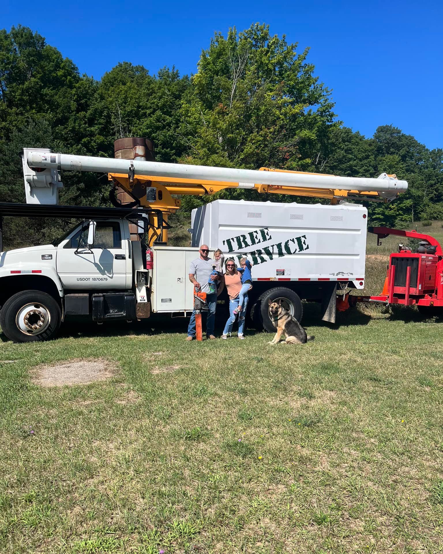 A family standing next to a utility truck with a bucket lift, labeled 'TREE SERVICE', on a grassy area with trees in the background under a clear blue sky.