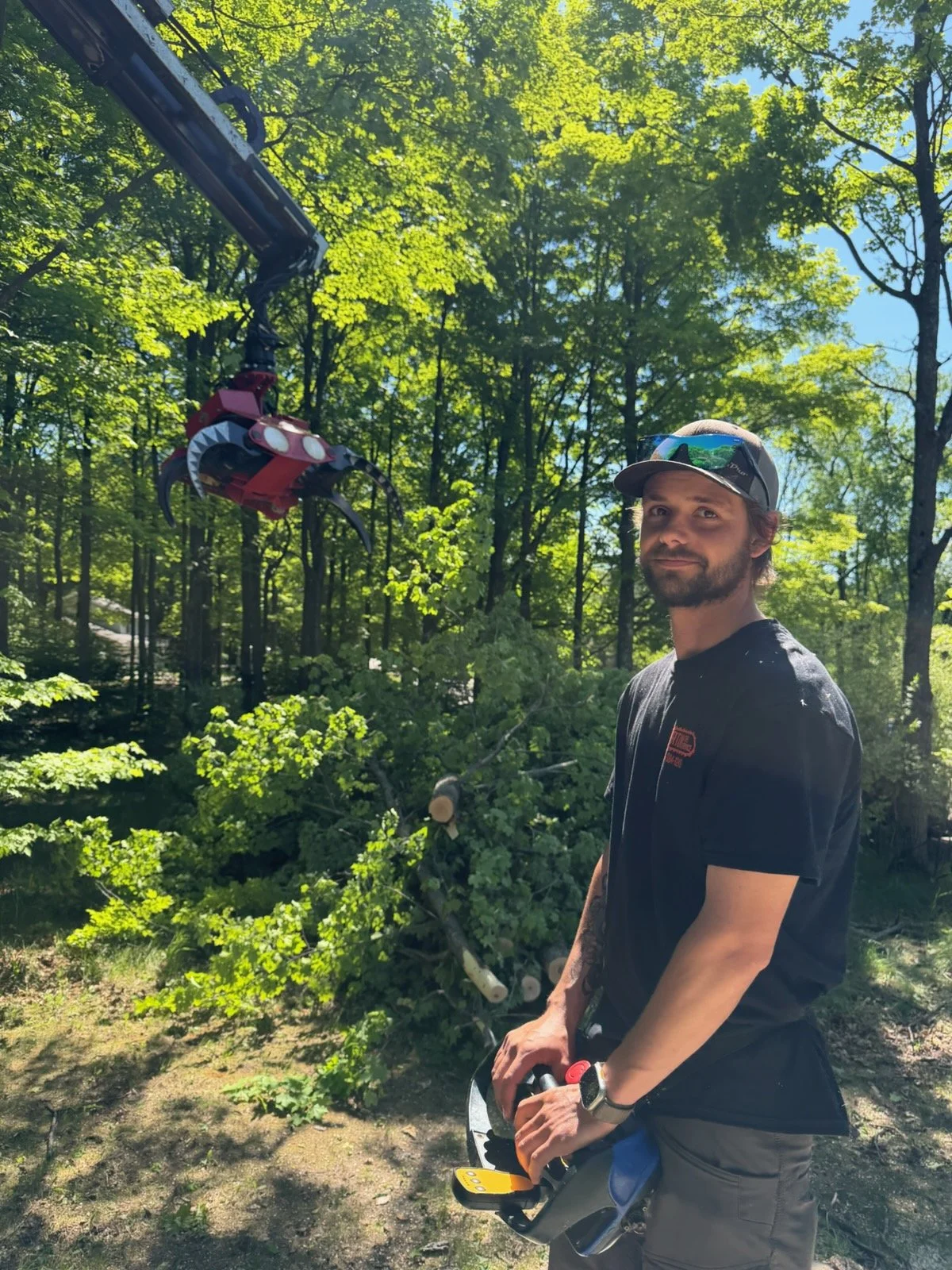 A smiling man standing outdoors in a wooded area, leaning against a tree with his hands in his pockets. He is wearing a light gray O'Brien garage LLC baseball cap, a green and gray hoodie, and tan pants. The background features dense green foliage and a stack of cut logs.