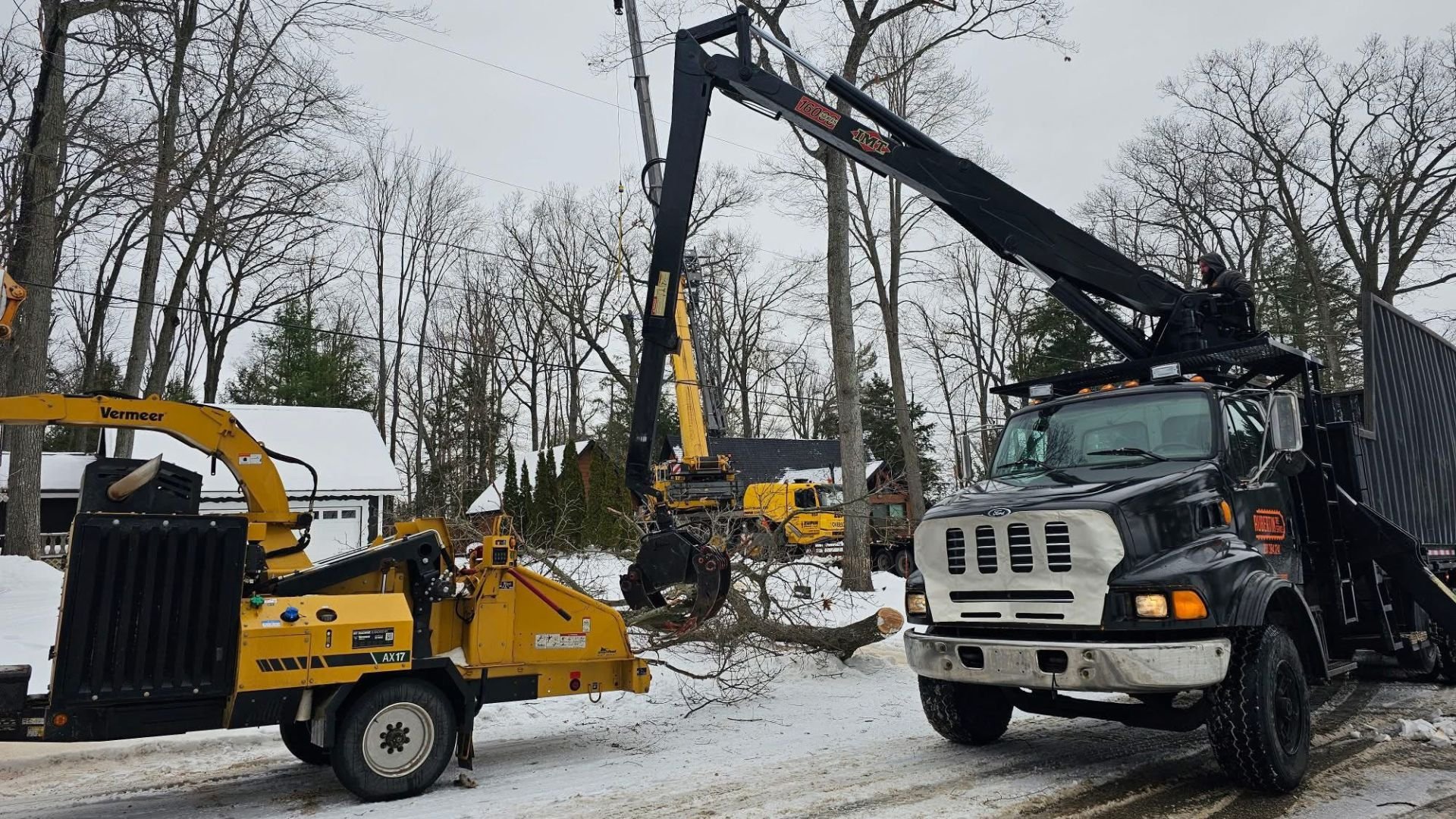 Tree trimming or removal work with a crane truck on a snowy residential street, with a background of leafless trees and houses.