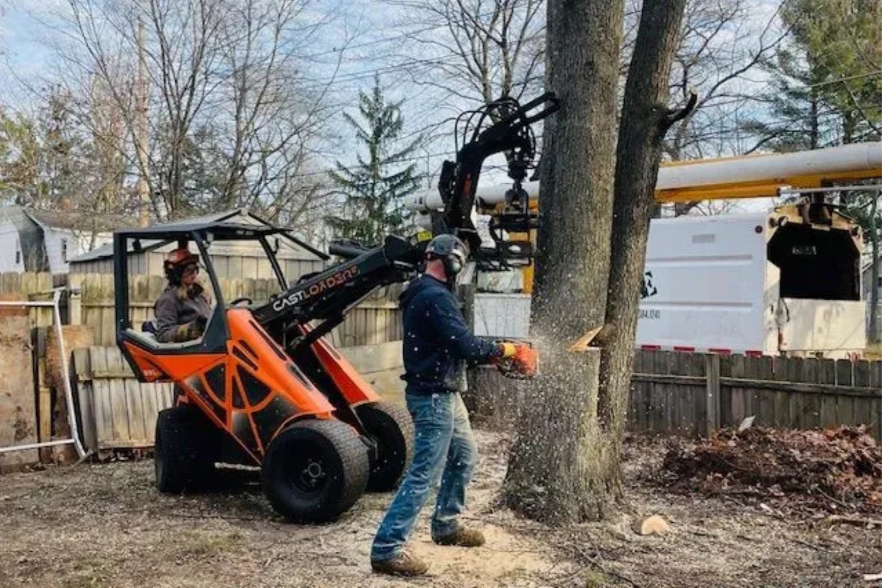 Two workers are cutting down a large tree in a backyard, using a chainsaw and a tree removal machine. One worker is operating the machine while the other is standing nearby holding a chainsaw, with wood debris visible on the ground.