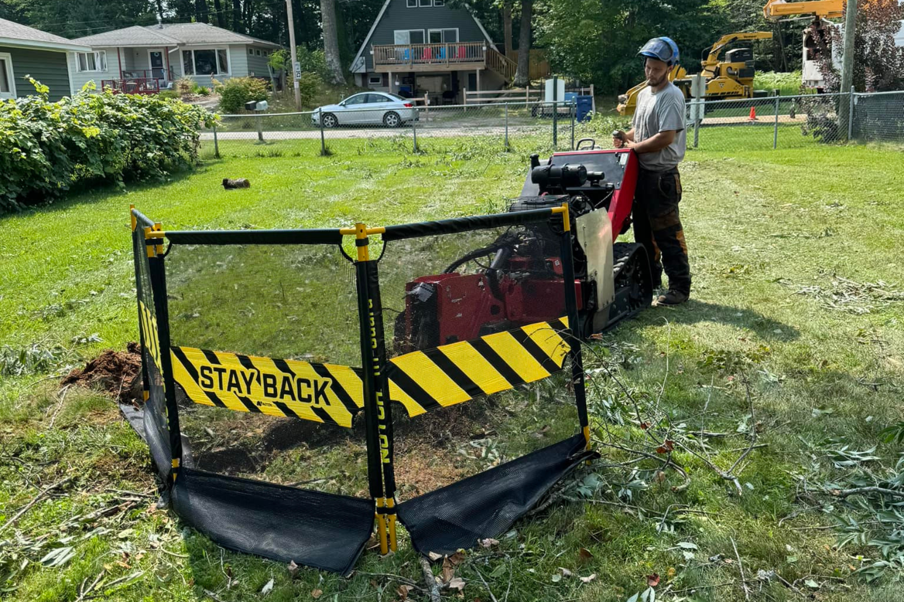 A man in a helmet operates a wood chipper in a backyard enclosed by a yellow and black caution barrier that reads 'STAY BACK.' The man is using a smartphone, and there are branches and debris around the chipper. Houses and a car are visible in the background.