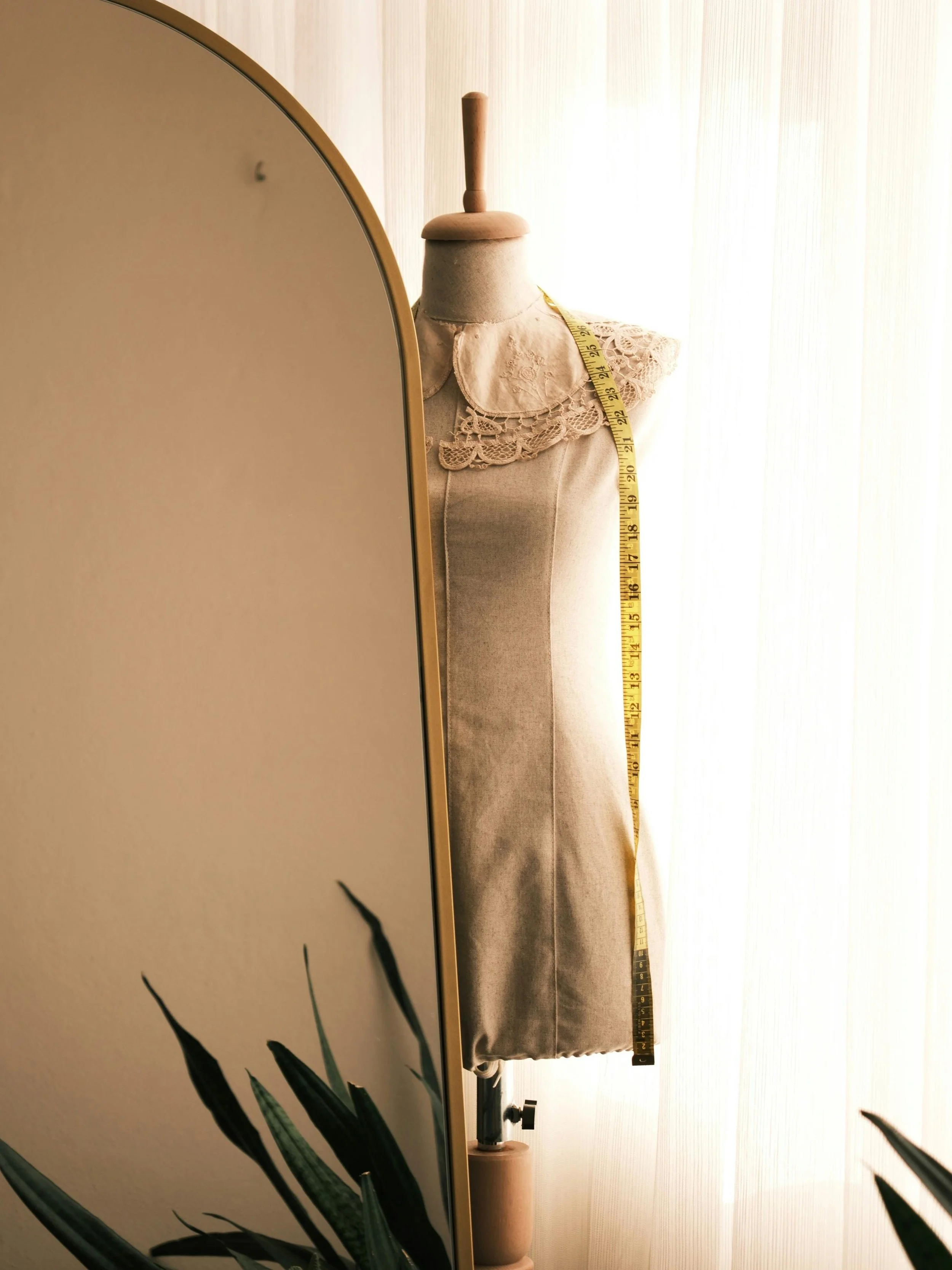 A dress form mannequin with a lace collar, a tape measure draped over it, and a wooden bottom stand. There is a mirror in the foreground and a plant with long, pointed leaves in the lower left corner. The background has bright sheer curtains.