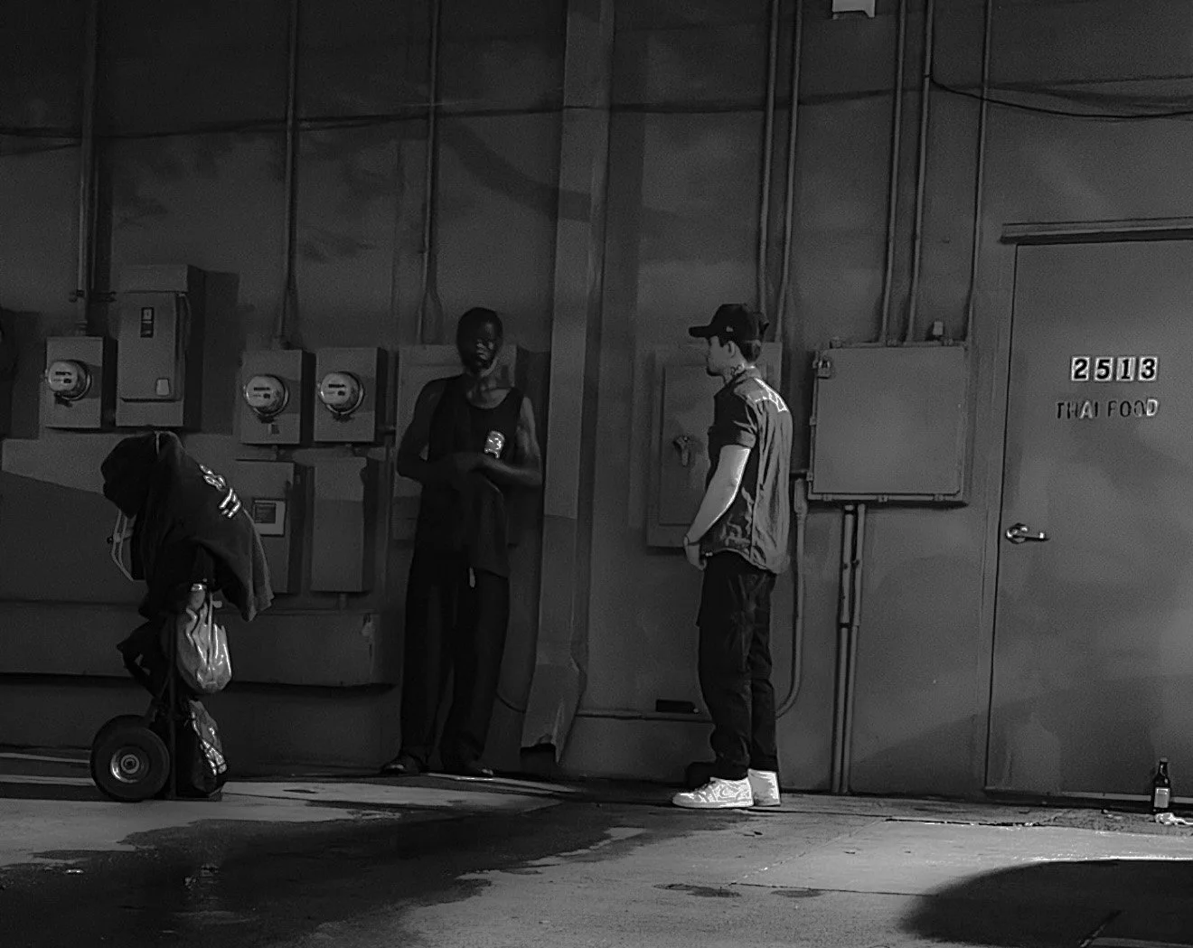Three people in an alleyway or loading dock area; one person with a backpack on a motorized skateboard, another person leaning against the wall with a backpack and wearing dark clothing, and a third person wearing a baseball cap and dark clothing standing near a door labeled '253 TRAY FOOD'; electrical panels and wires on the wall.