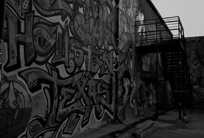 A black-and-white photo of a graffiti-covered wall with a metal fire escape staircase on the right.