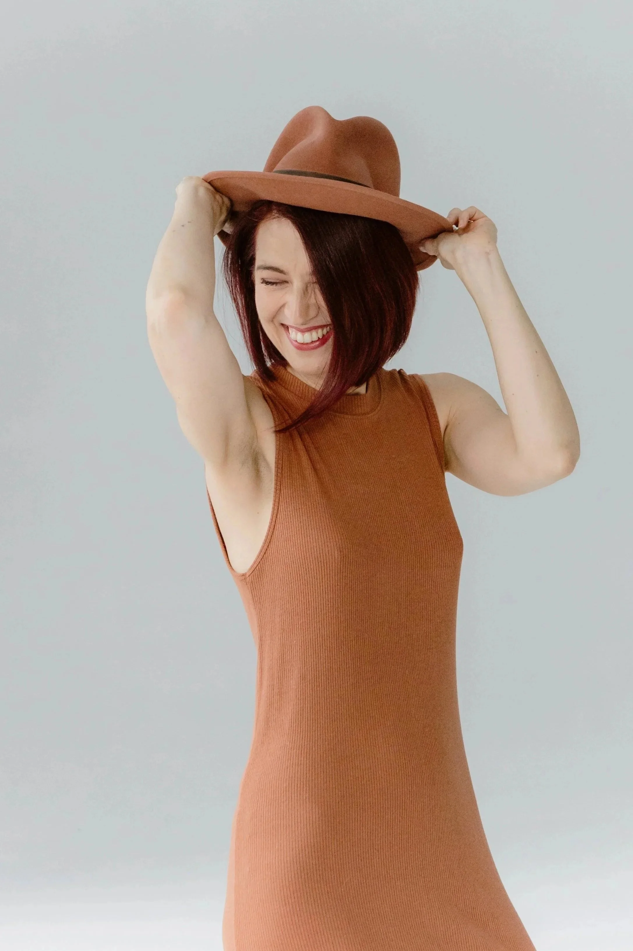 A woman with red hair smiling and holding a brown hat on her head, wearing a sleeveless orange top, standing against a plain light background.
