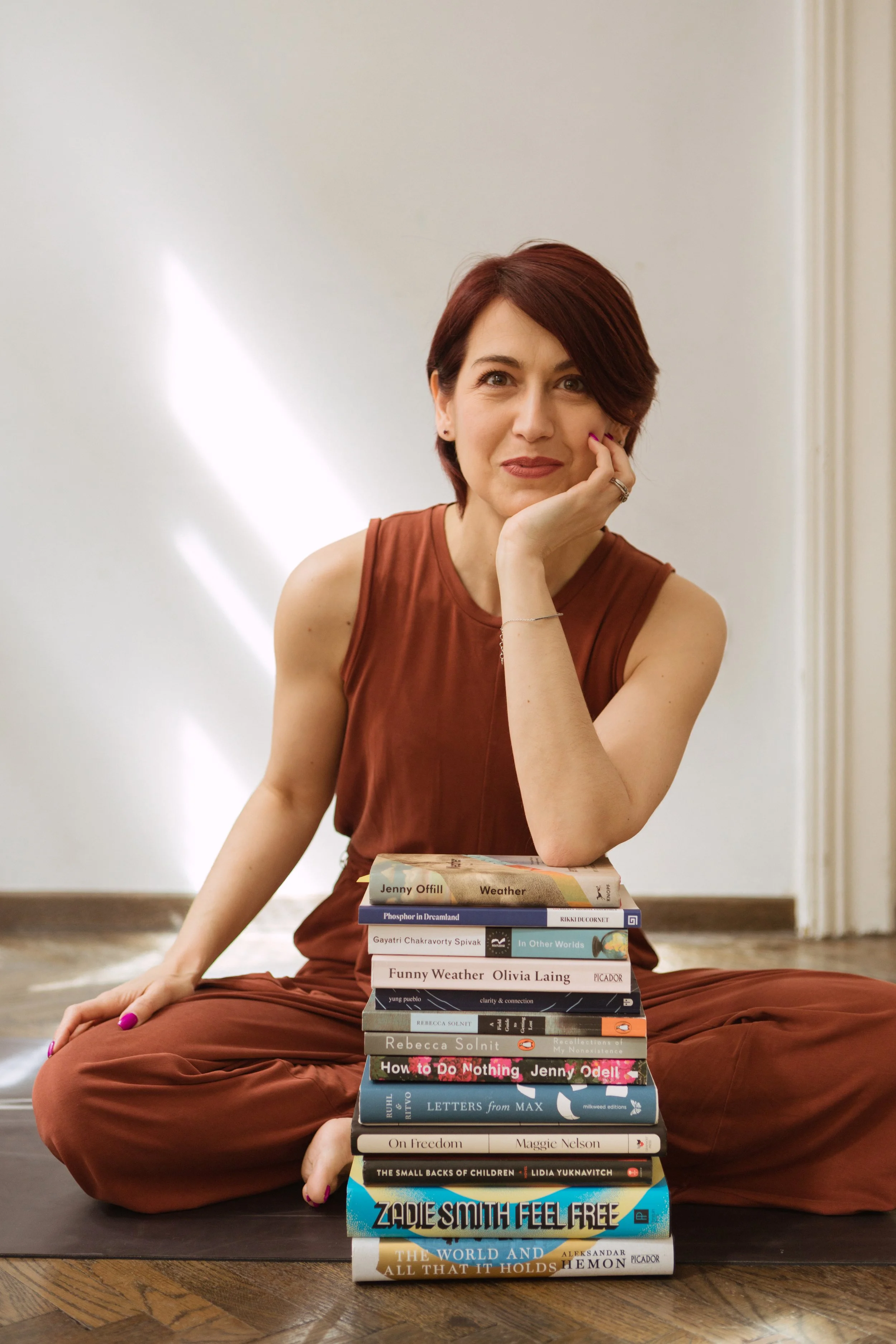 A woman with short red hair sitting on the floor, barefoot, in front of a stack of books, resting her chin on her hand, with a relaxed and thoughtful expression.
