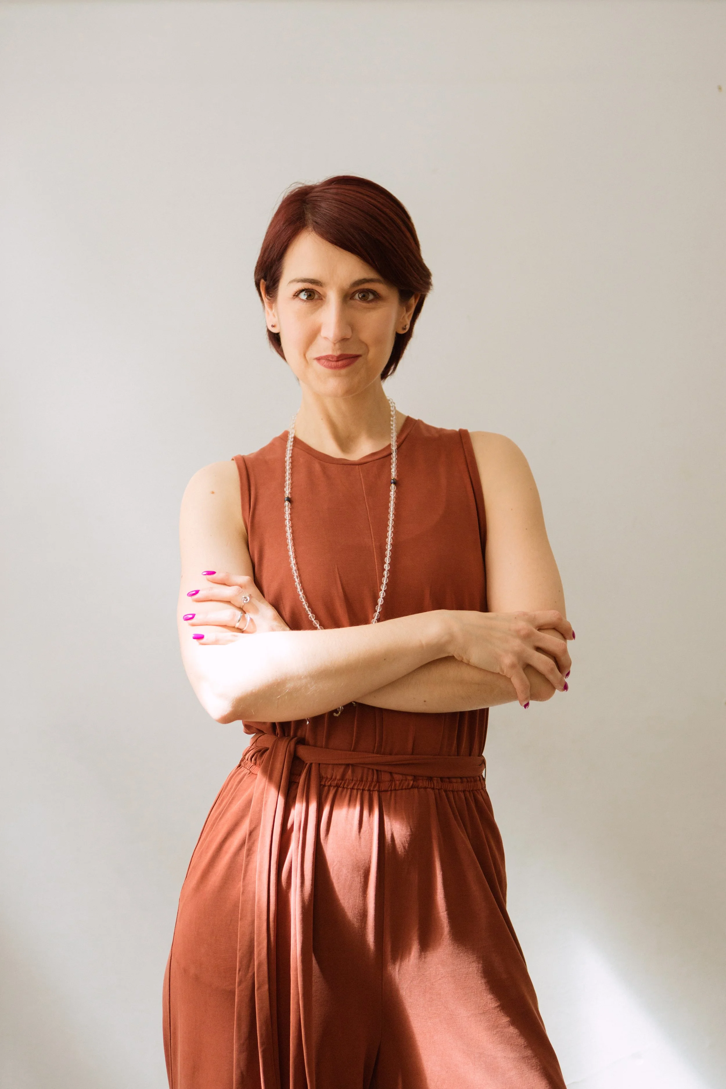 A woman with short auburn hair, wearing a rust-colored sleeveless top and matching pleated pants, standing with her arms crossed in front of a plain, light-colored wall.