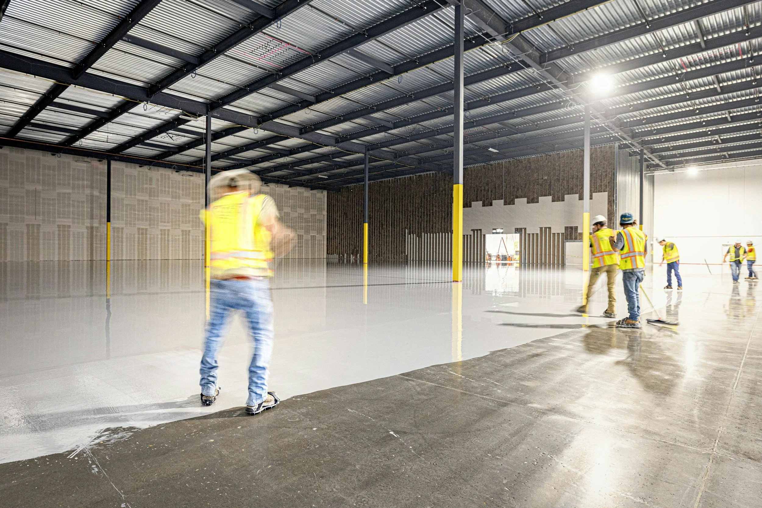 Construction workers in safety vests and helmets skating inside a large indoor industrial space, possibly a warehouse or factory under construction.