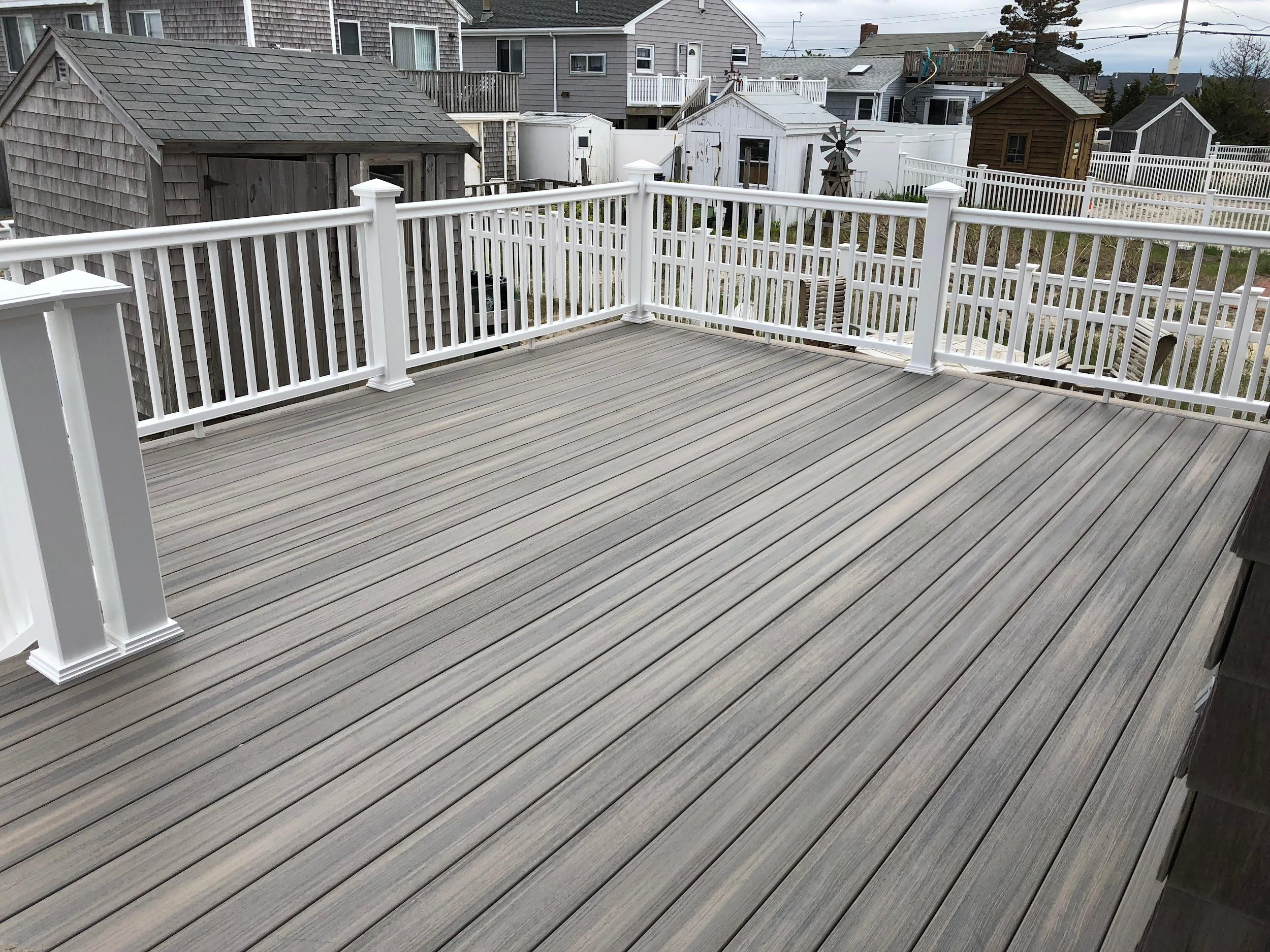 An outdoor wooden deck with white railing, overlooking neighboring houses with various small storage sheds and a windmill decoration in a residential area.  Mastercraft Building & Remodeling offers exterior construction services including decks.