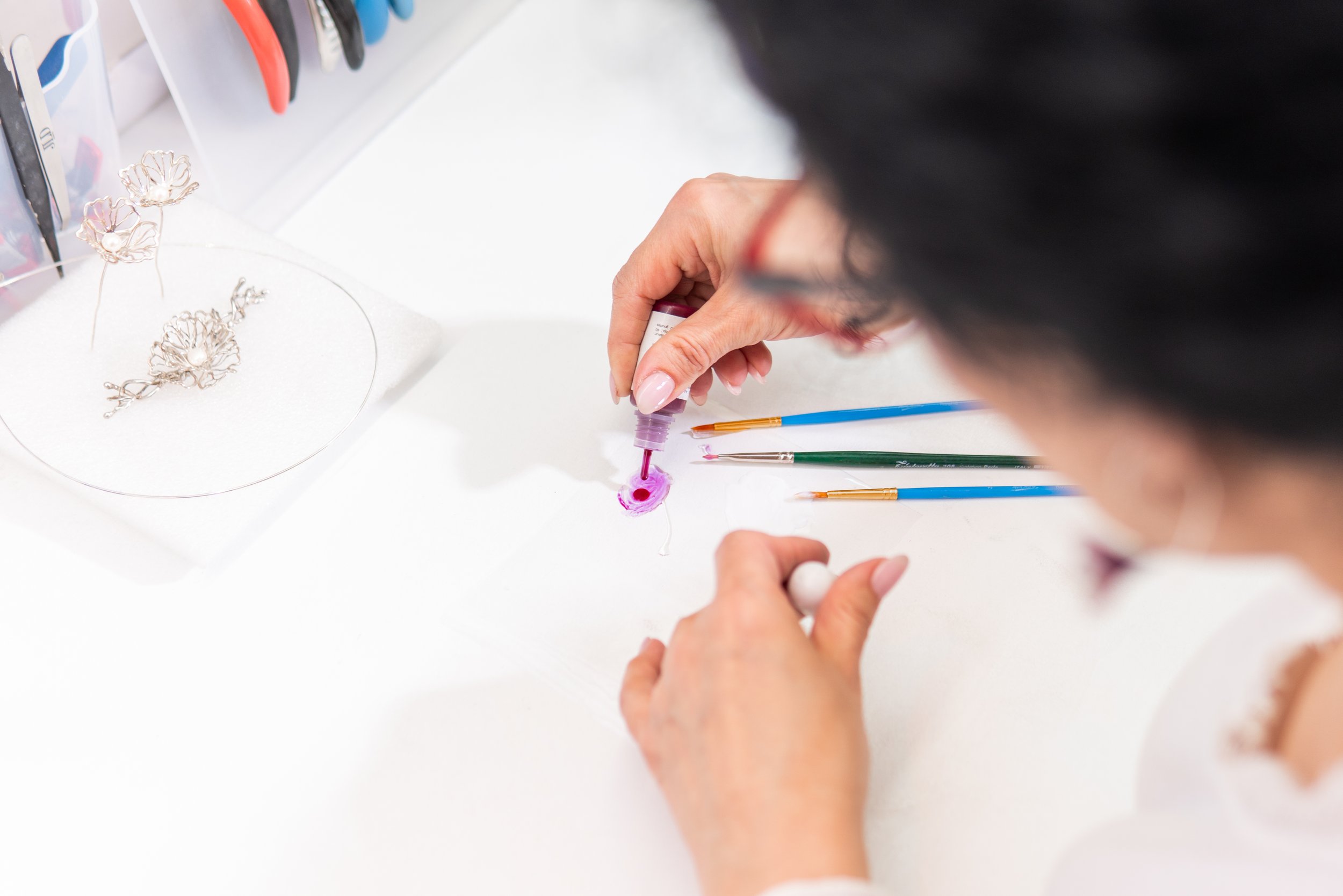 A person with black hair and glasses is using a paintbrush and a dropper to create jewelry, with jewelry pieces on a white surface and color tools around.