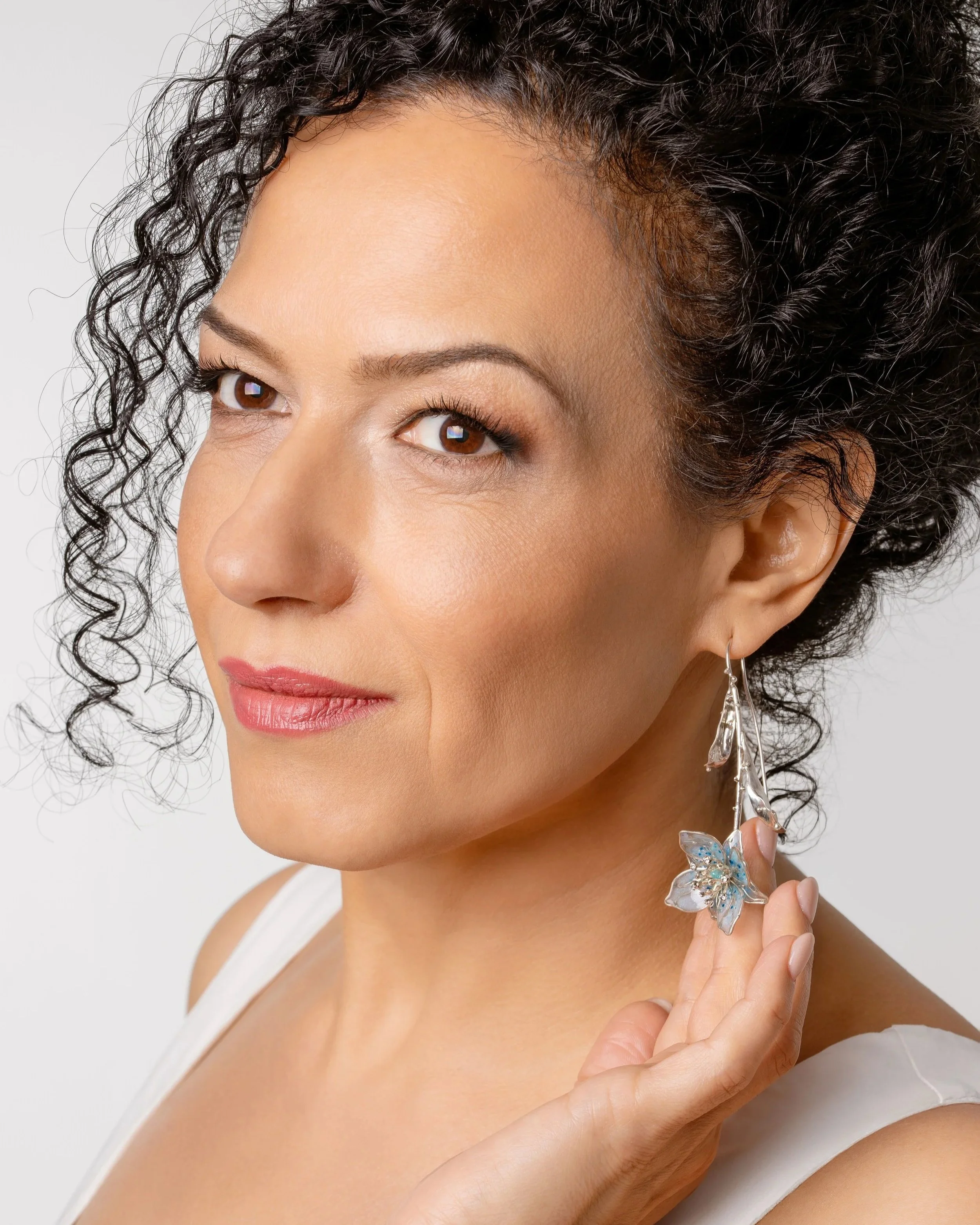 A woman with curly black hair holding a butterfly-shaped earring near her face. She has brown eyes, well-groomed eyebrows, and smooth skin. She is wearing a white top and has minimal makeup.
