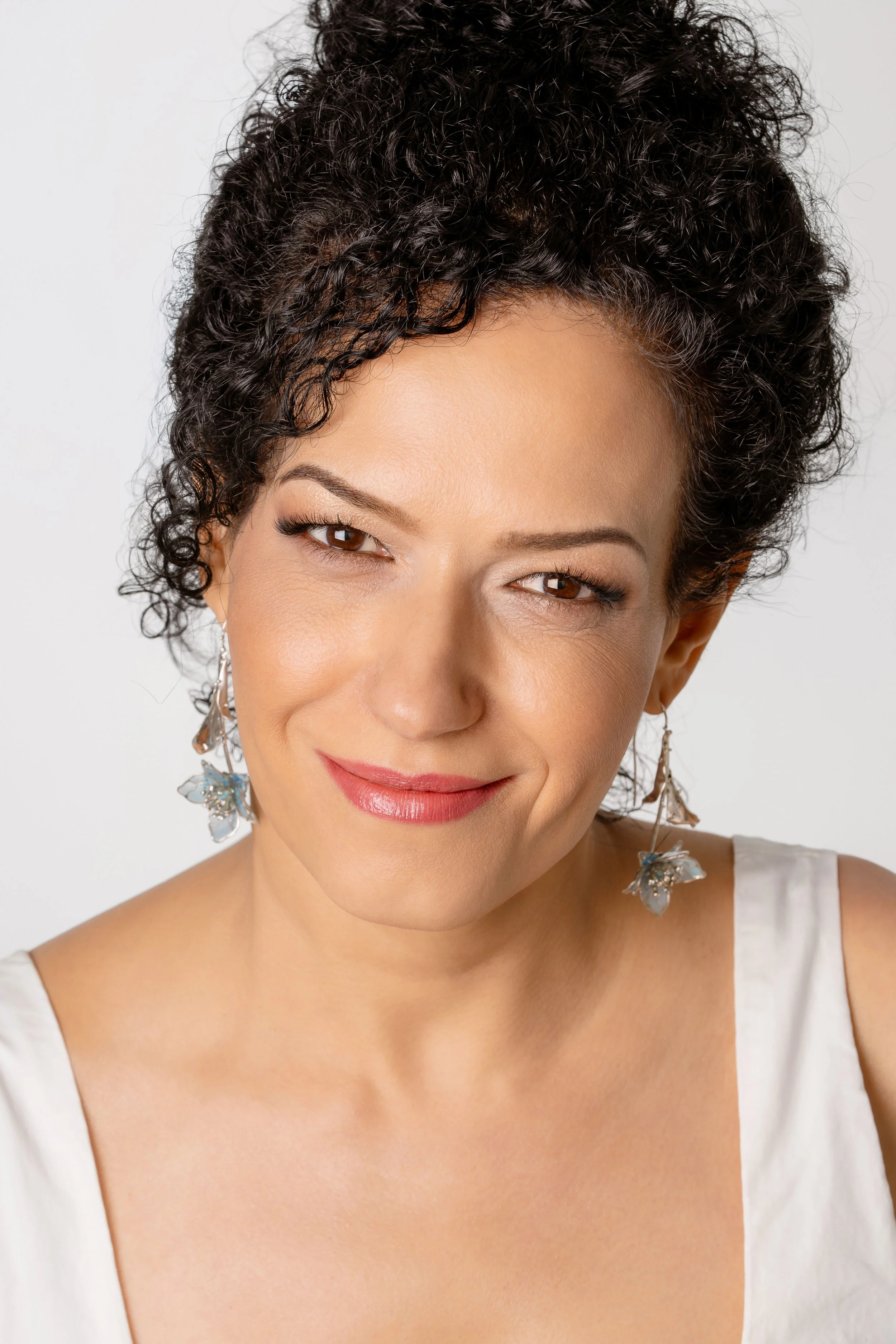 A woman with short dark curly hair, wearing earrings with butterfly and leaf designs, smiling, dressed in a white sleeveless top, against a plain background.