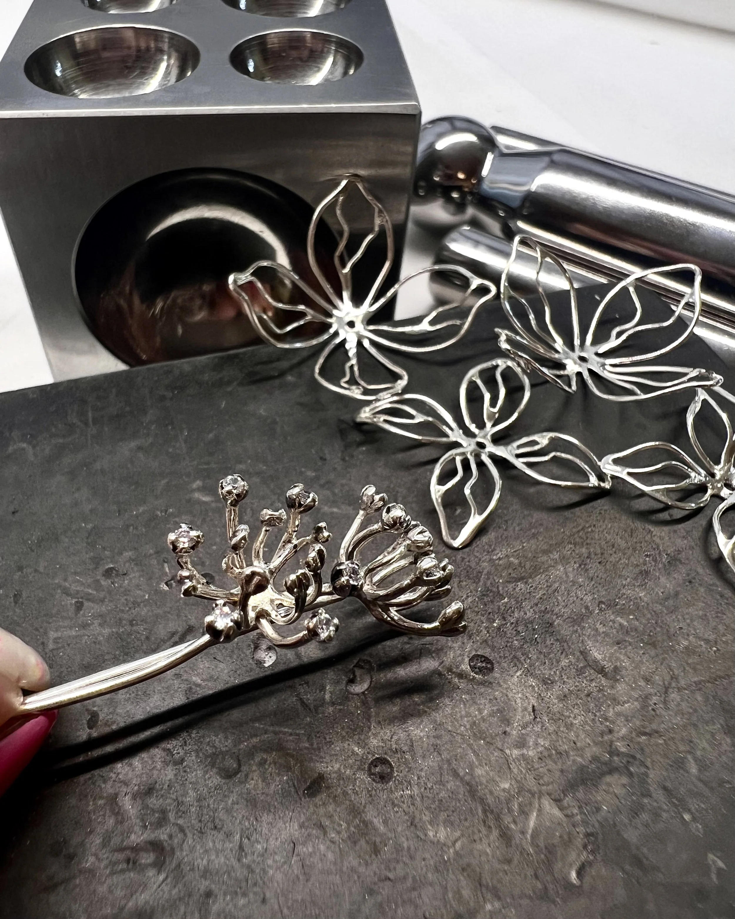 Silver earrings shaped like butterflies and a decorative flower, on a dark surface with a metal object and a jewelry-making tool in the background.