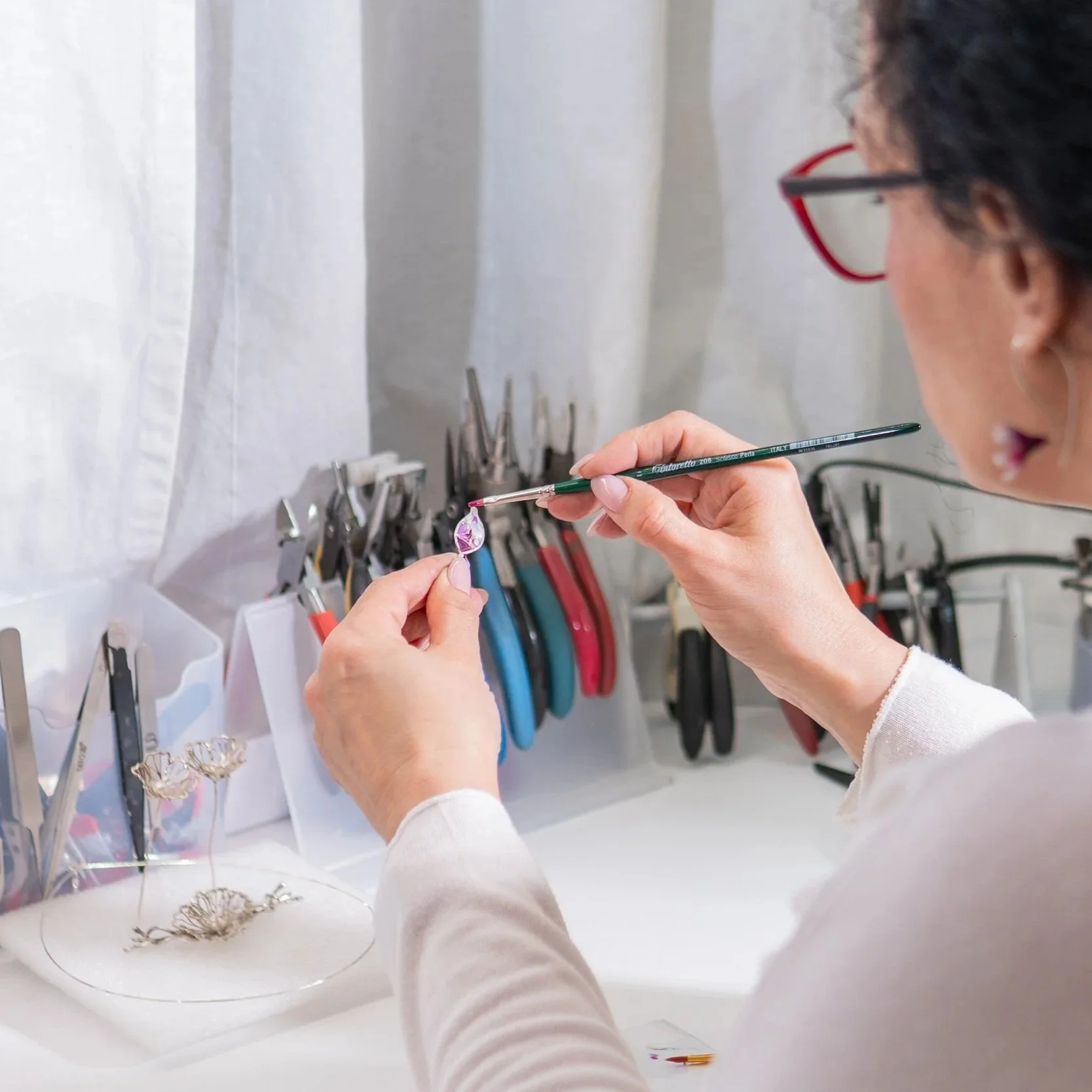 Person working on jewelry making, holding a small object with a paintbrush, in a workspace with tools and jewelry pieces on display.