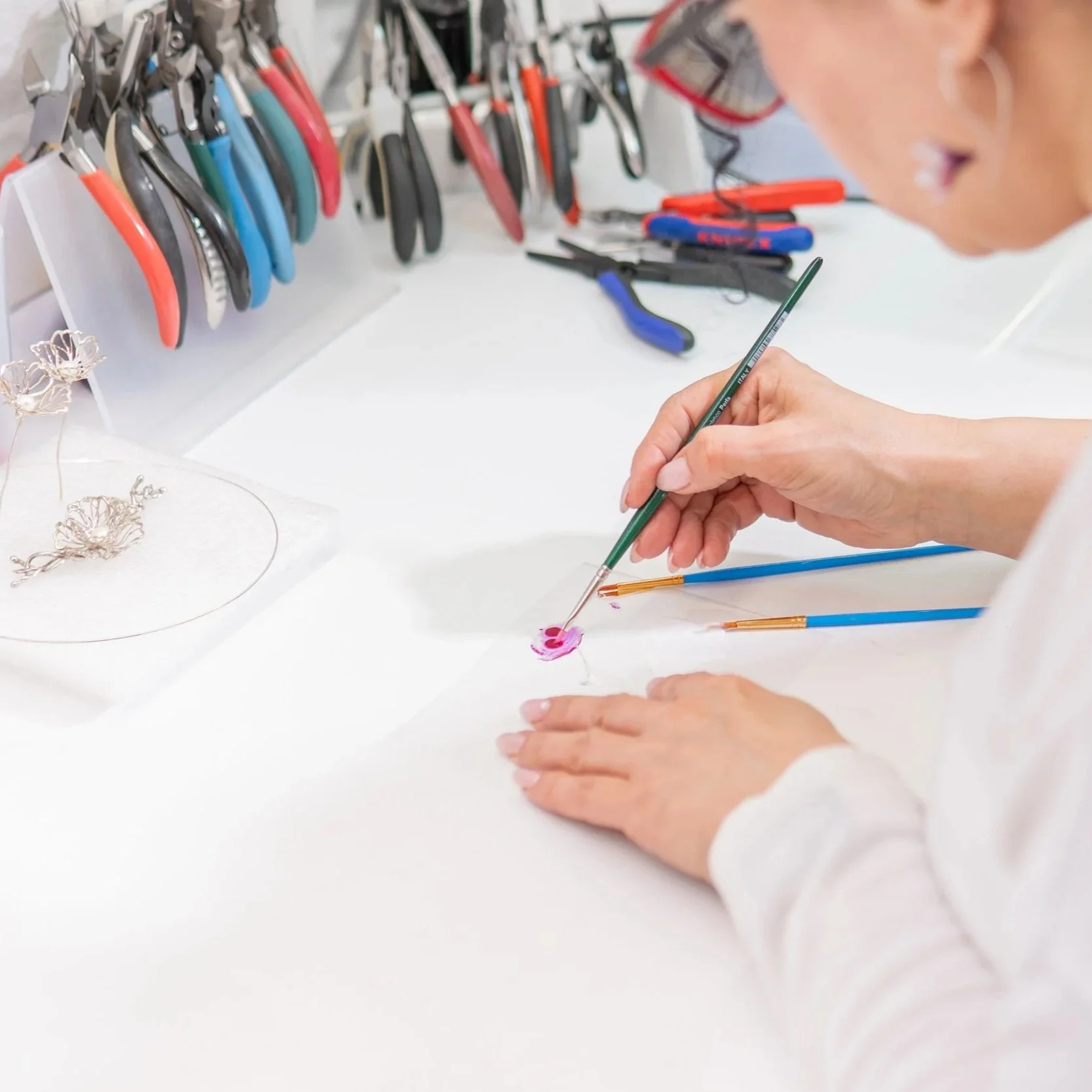 Person with glasses painting a pink flower on paper with a thin paintbrush, on a white table with various tools and jewelry.