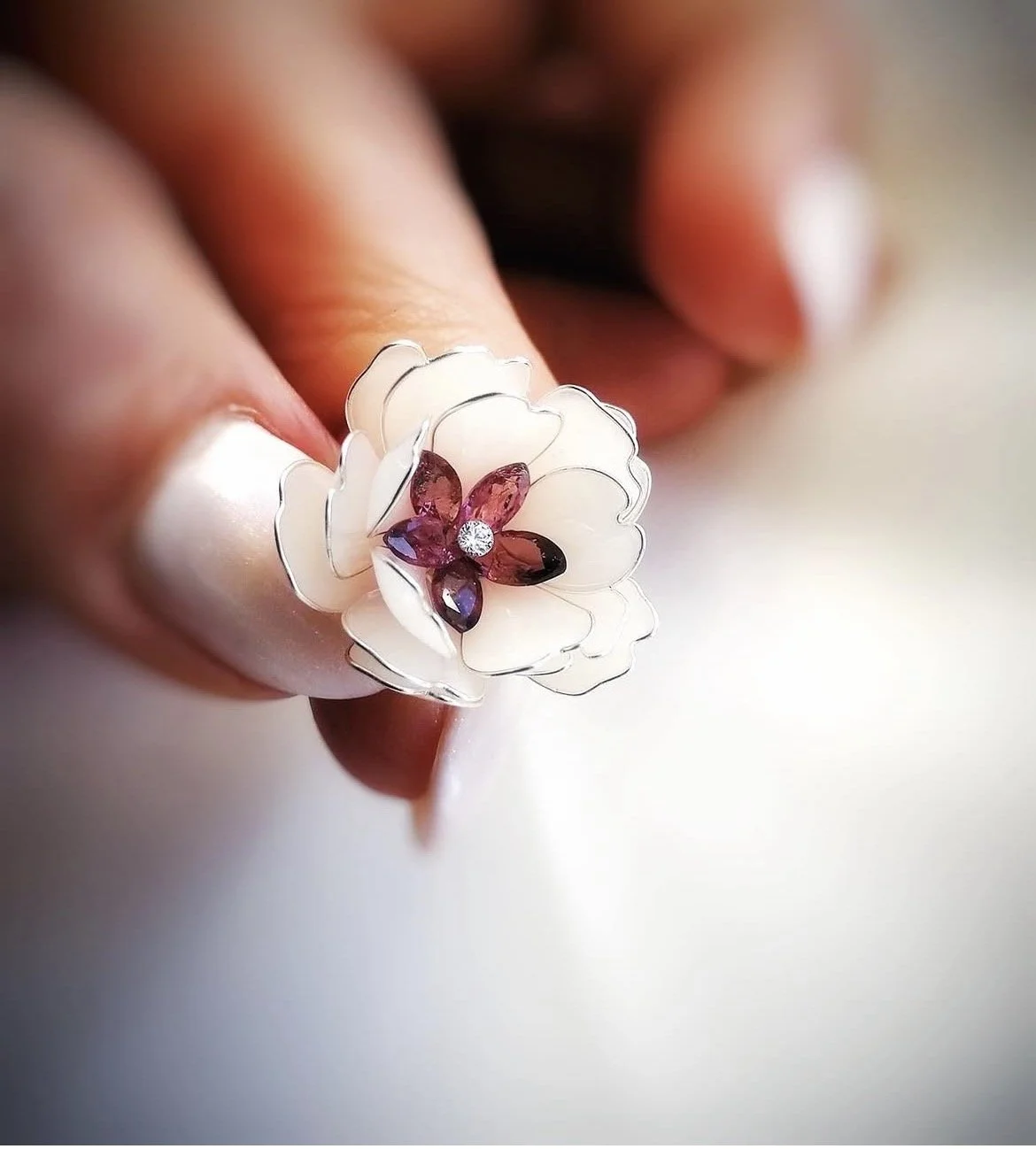 Close-up of a flower-shaped ring with a white petals design, featuring purple and black gemstones at the center and a small diamond accent, worn on a person's finger.