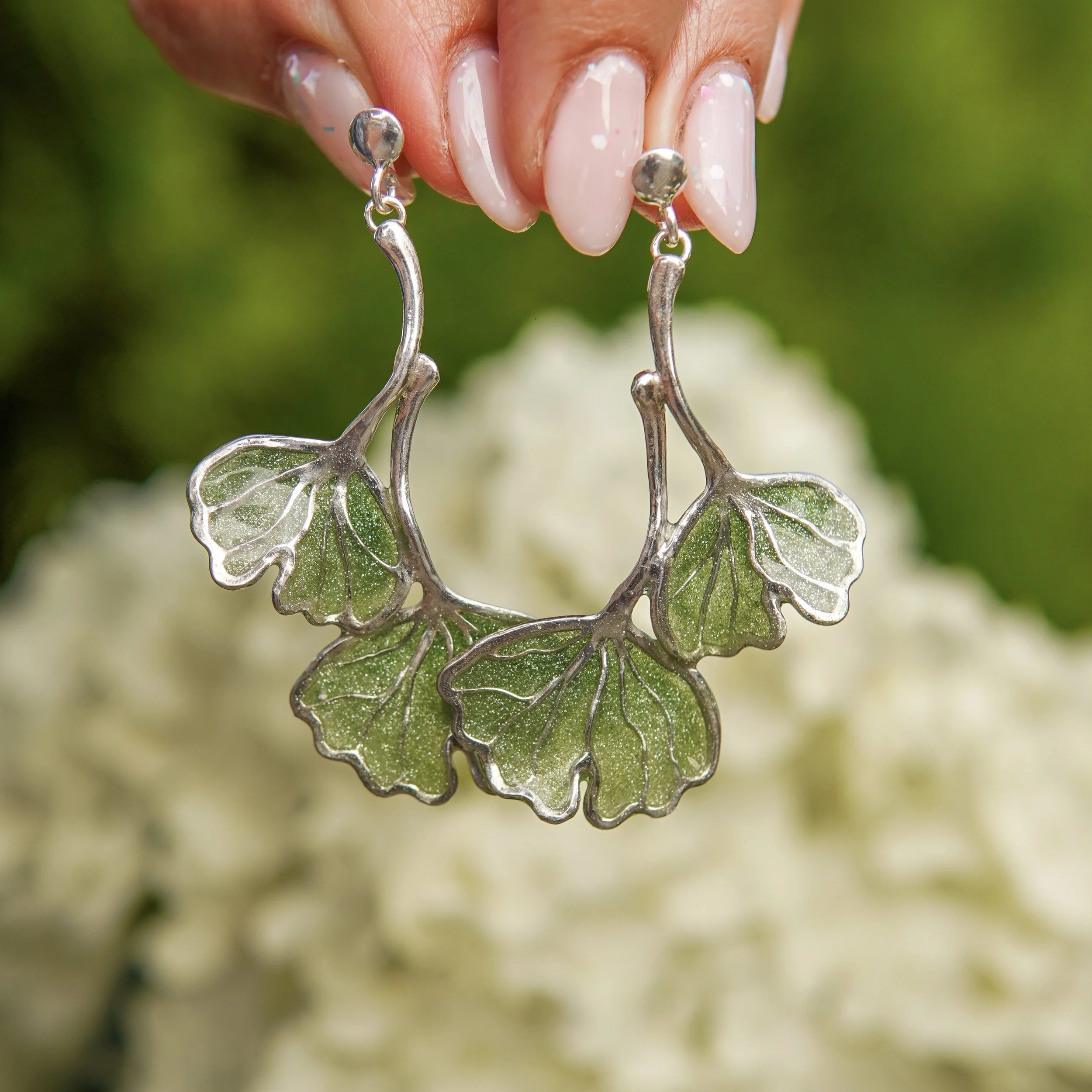 Close-up of metallic earrings with leaf shapes, held between fingers with painted nails, blurred green background.