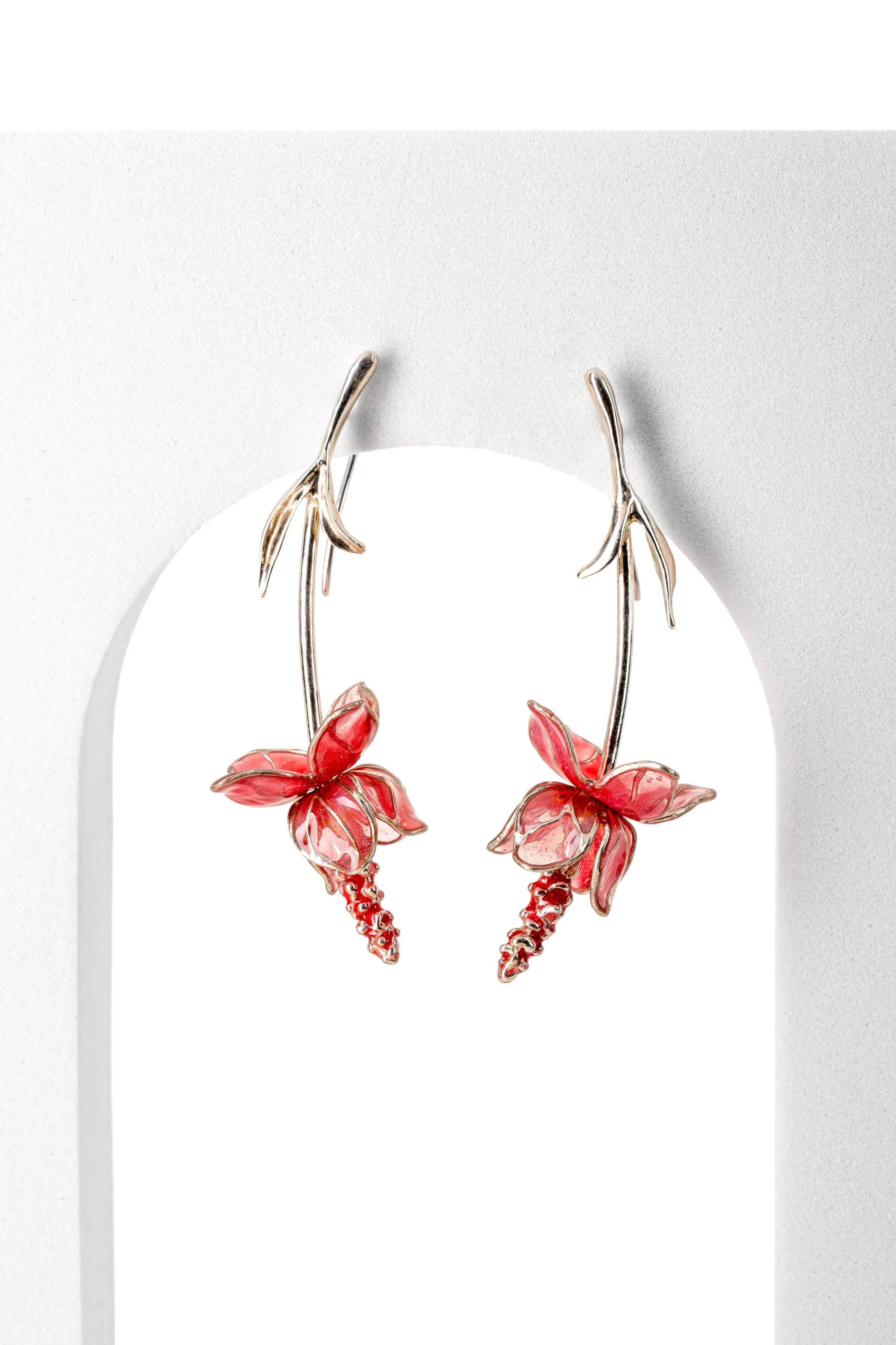 Silver earrings designed as red flowers with detailed petals and dangling chains, hanging on a white display stand.