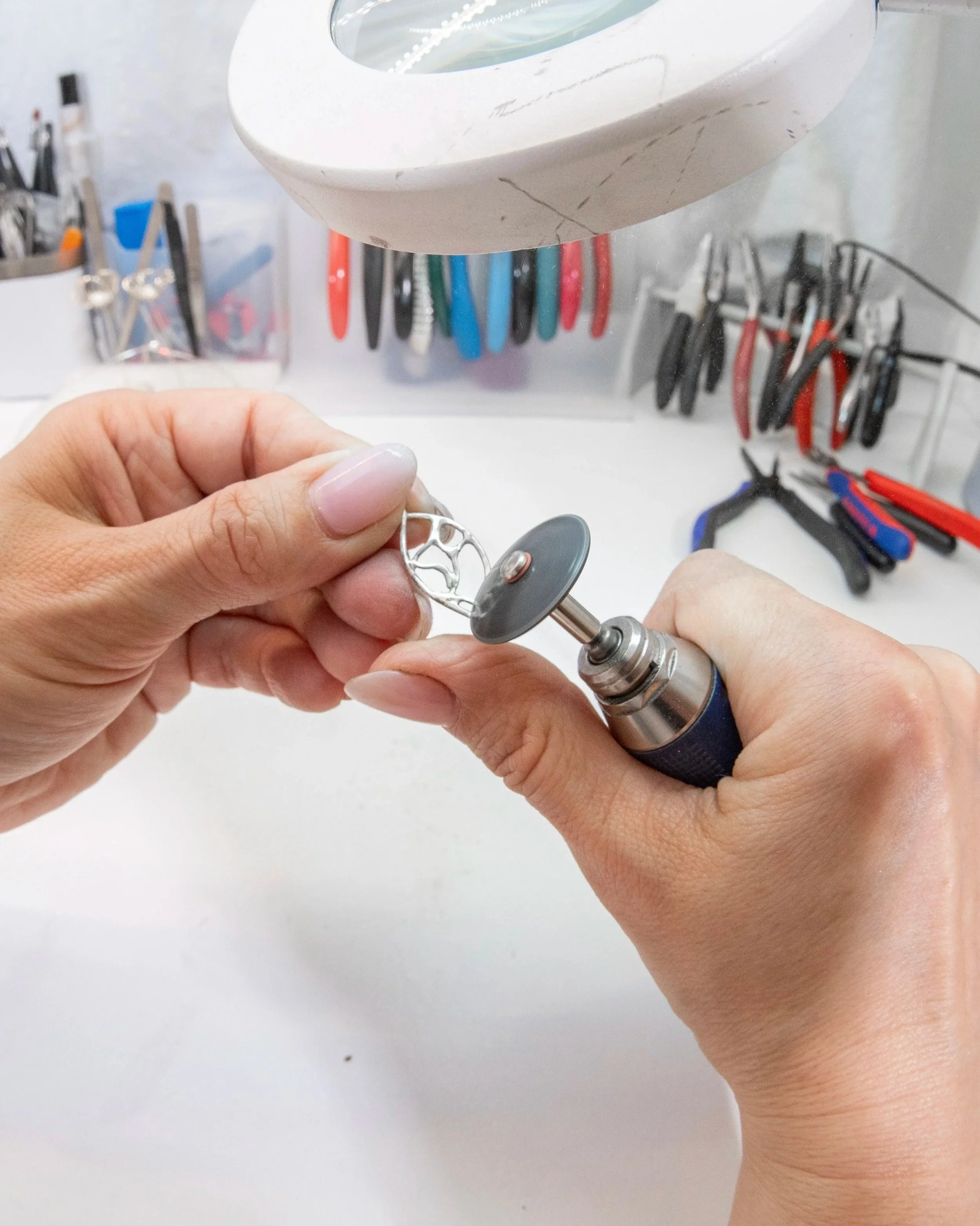 Person polishing a silver pendant with a rotary tool. The background shows various tools and jewelry-making supplies on a workbench.