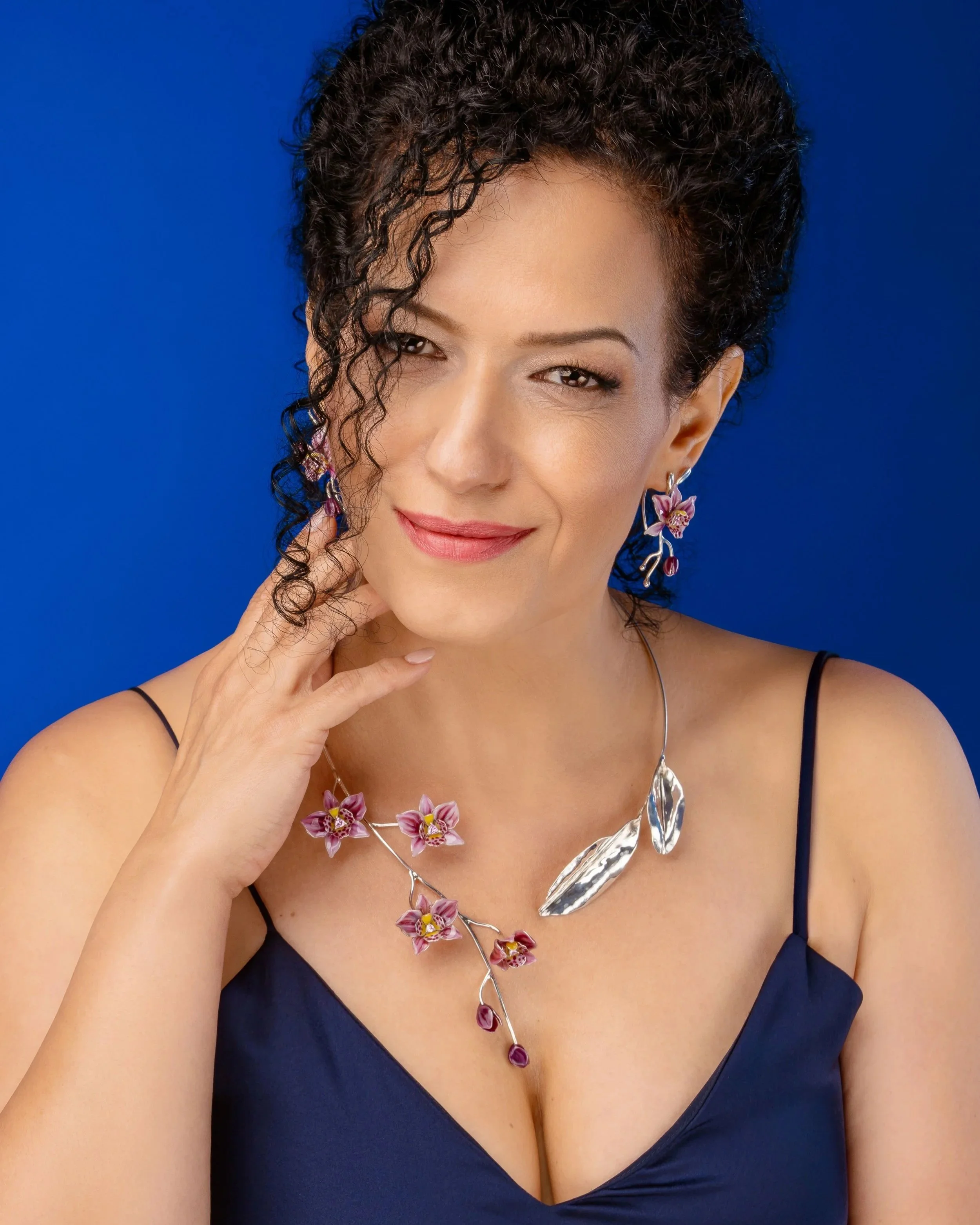 A woman with curly dark hair wearing a dark blue dress and floral jewelry, including a necklace and earrings, against a solid blue background.