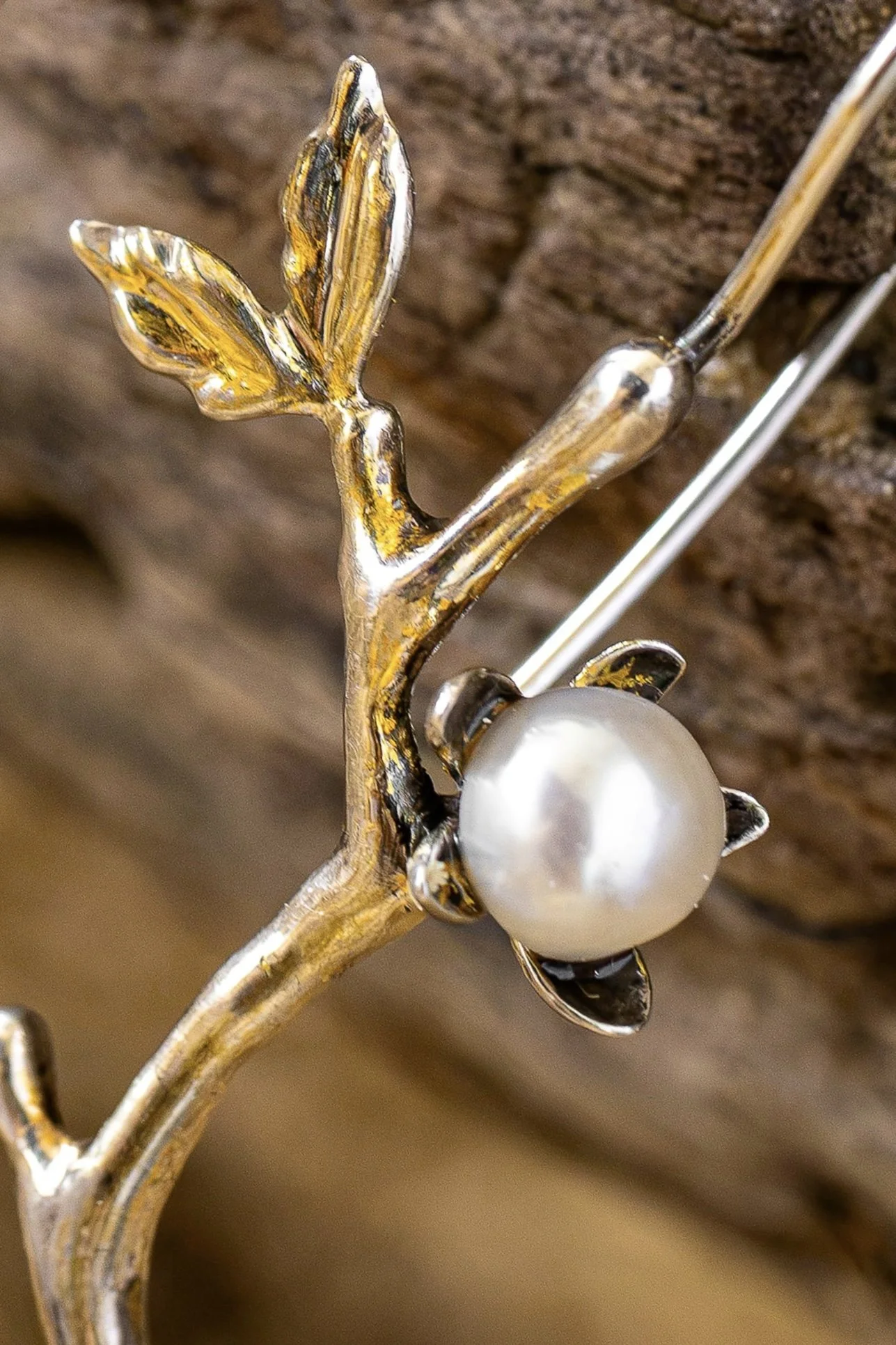 Close-up of a decorative jewelry piece resembling a branch with a pearl at the base, gold-colored leaves, and black accents, set against a blurred wooden background.