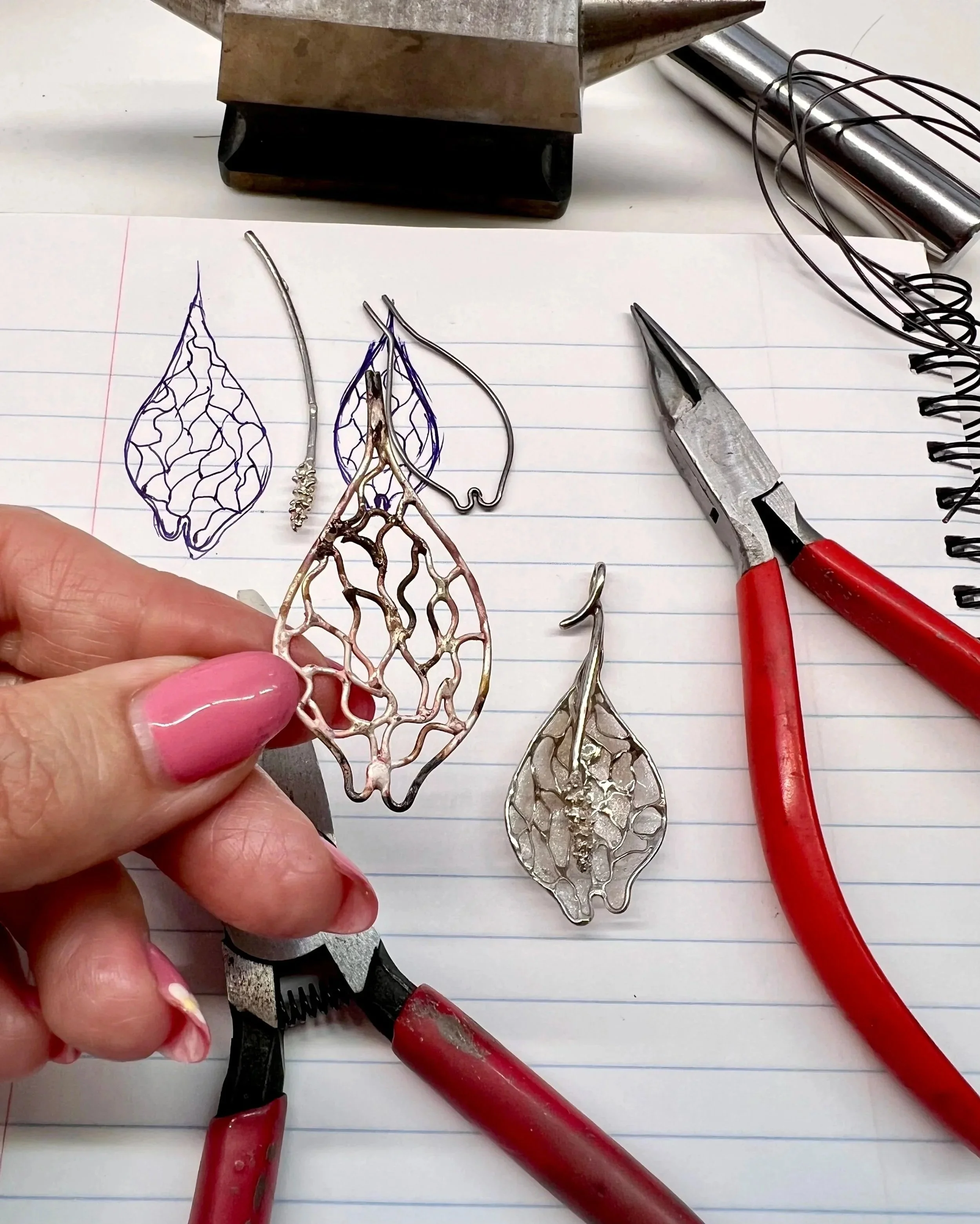 A person with pink nail polish holds a metal leaf-shaped jewelry piece over lined notebook paper. Various wire and metal jewelry components, pliers, and tools are on the paper, including a pair of red-handled pliers, a coil of wire, and partial metal substrucures.