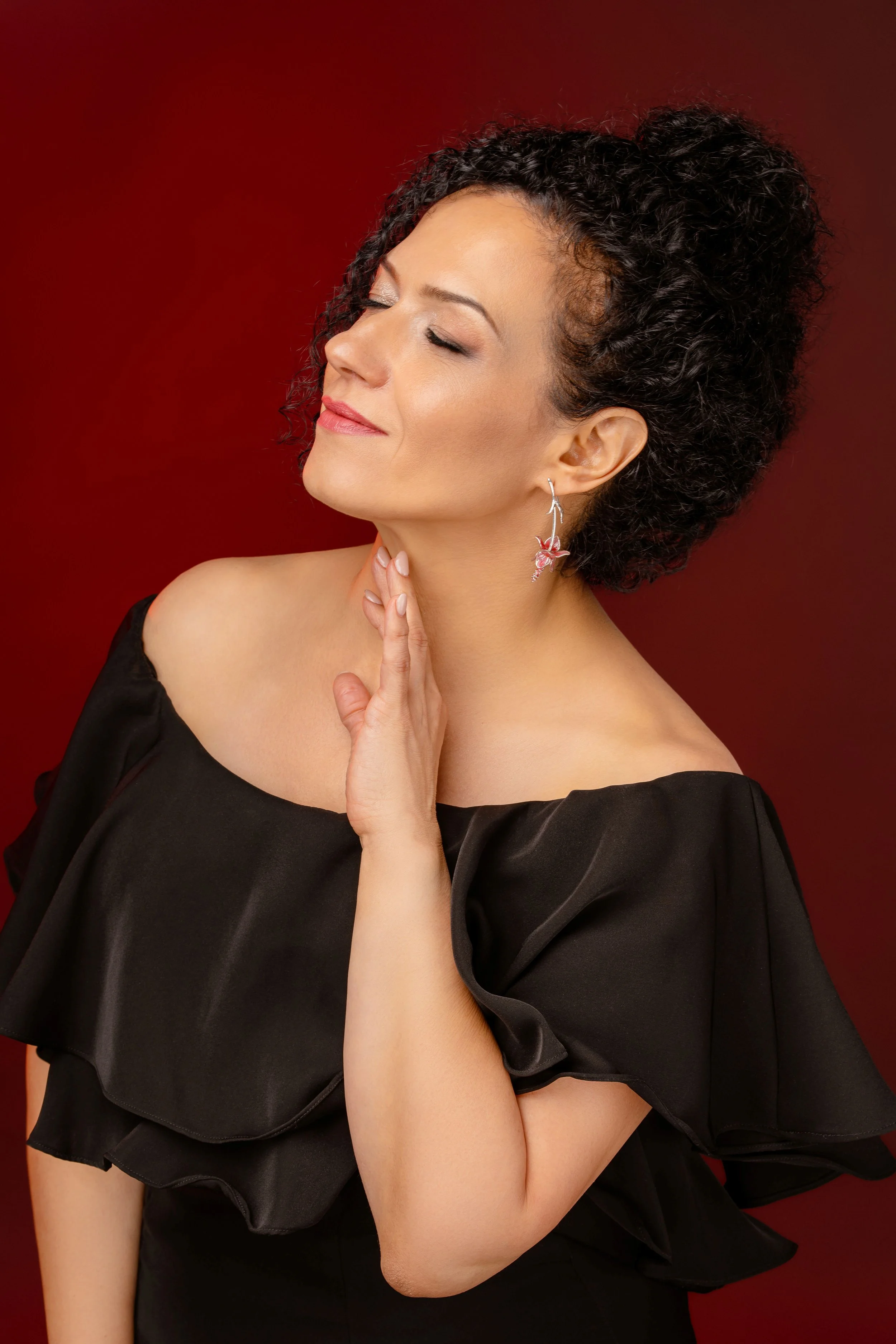 A woman with short, curly black hair and fair skin, posing with her eyes closed and a gentle smile, wearing a black off-the-shoulder dress with ruffled sleeves, an earring shaped like a flower, and standing against a red background.