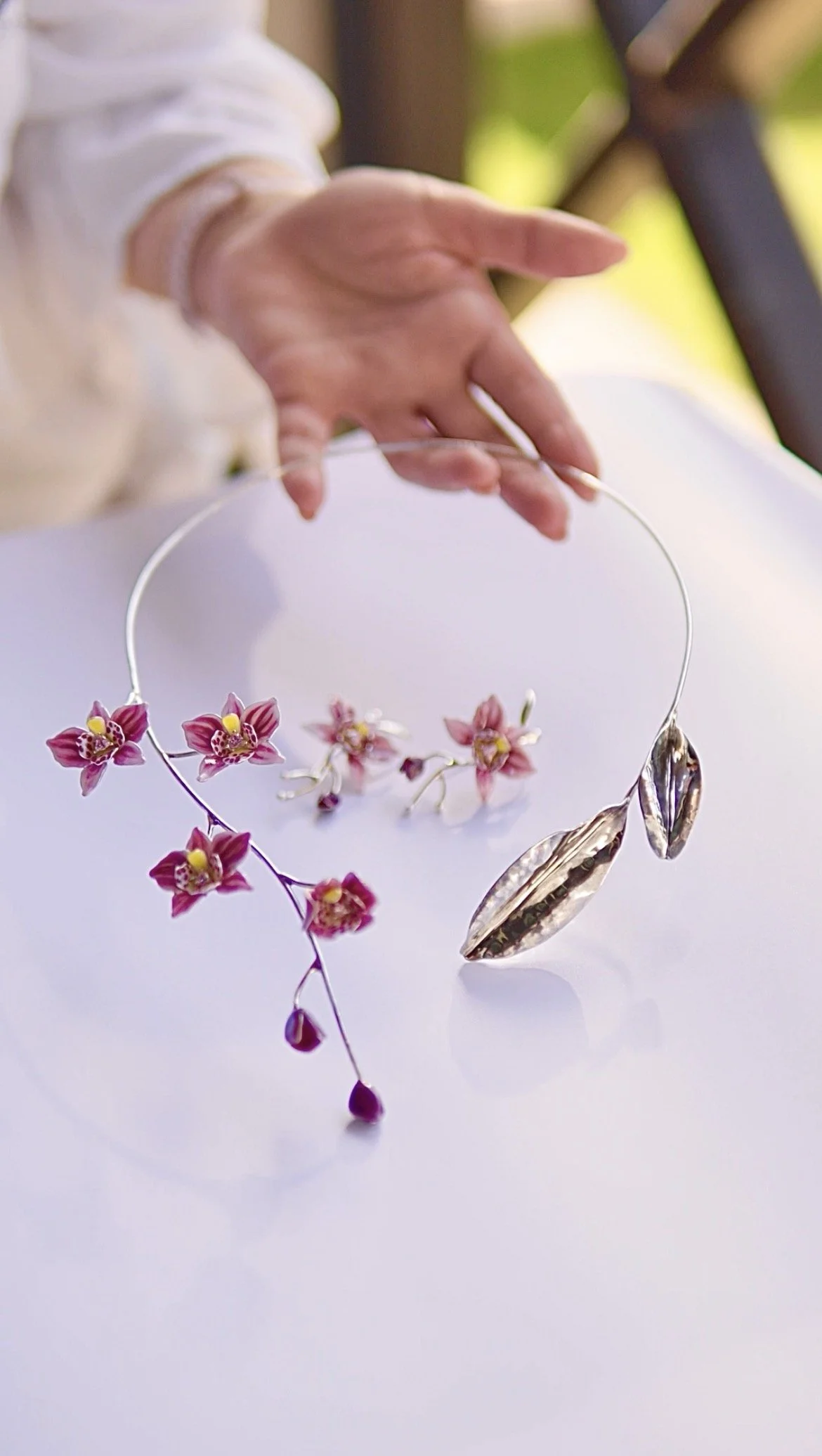 A close-up of a person holding a silver floral headpiece with pink orchids and metallic leaves, resting on a white surface.