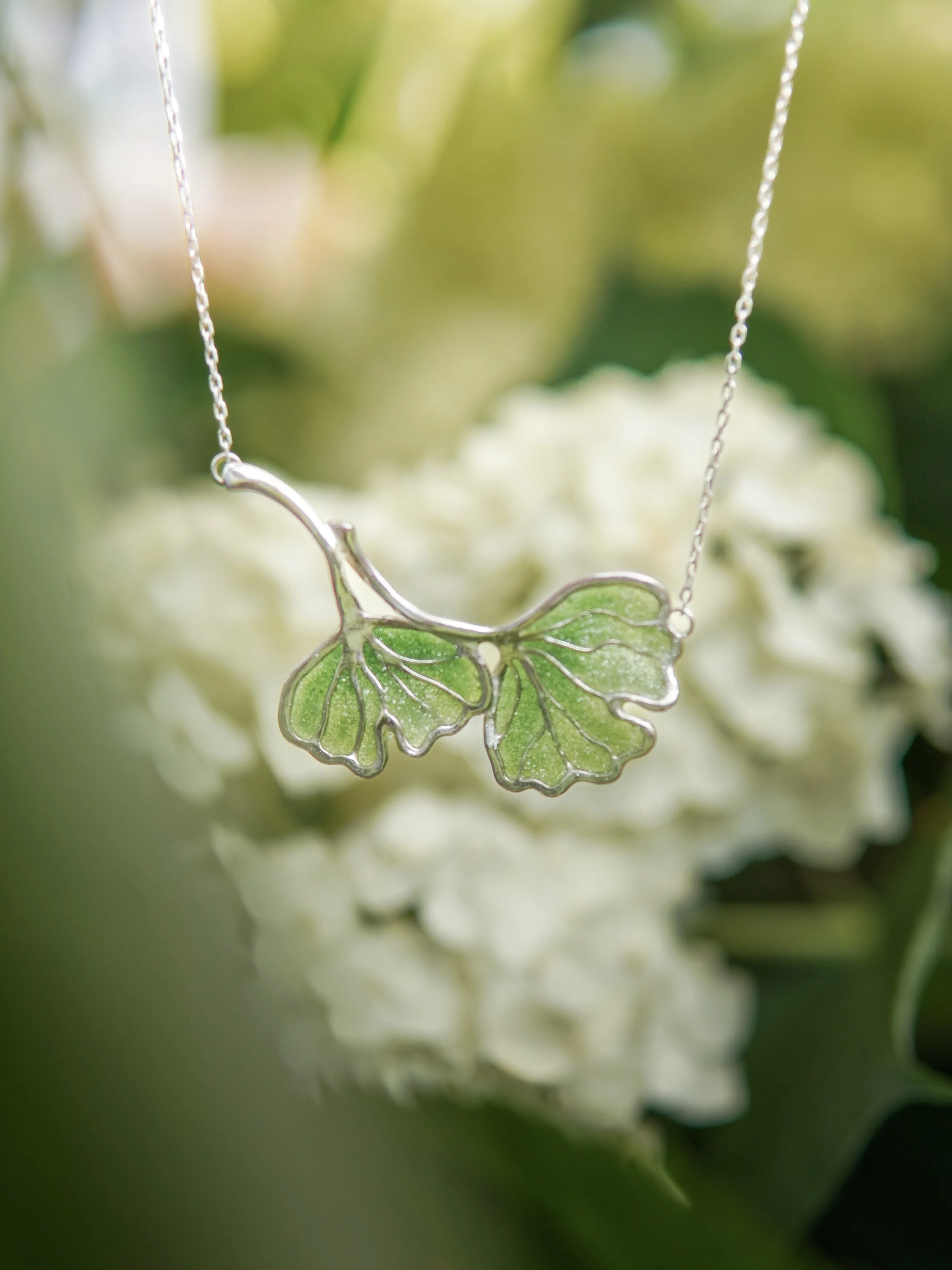 Close-up of a necklace pendant shaped like a green leaf with silver outlining and chain, with blurred white flowers and green background.