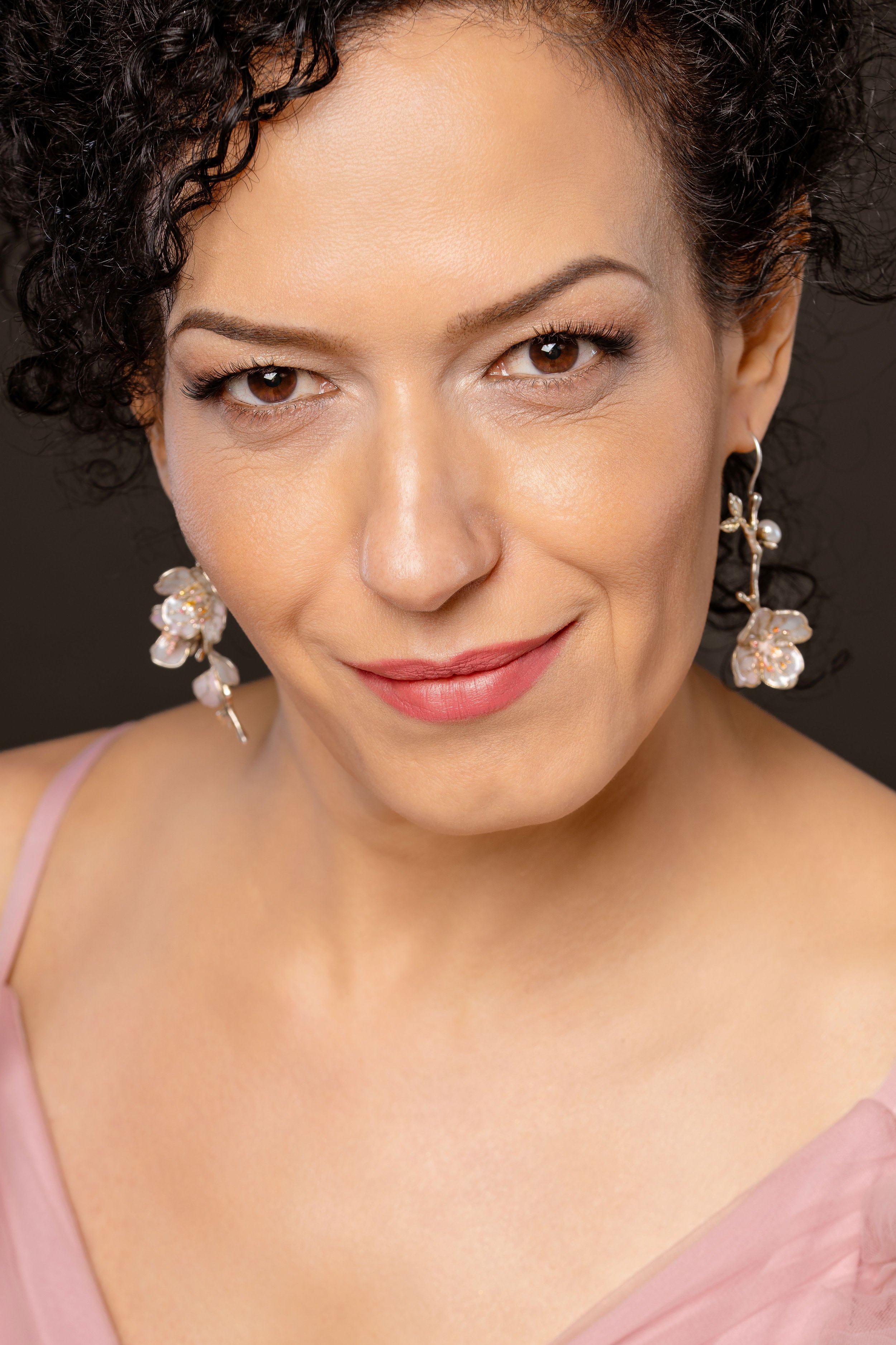 Close-up of a woman with fair skin, dark curly hair, brown eyes, and floral earrings, wearing a pink top with thin straps, smiling softly against a neutral background.