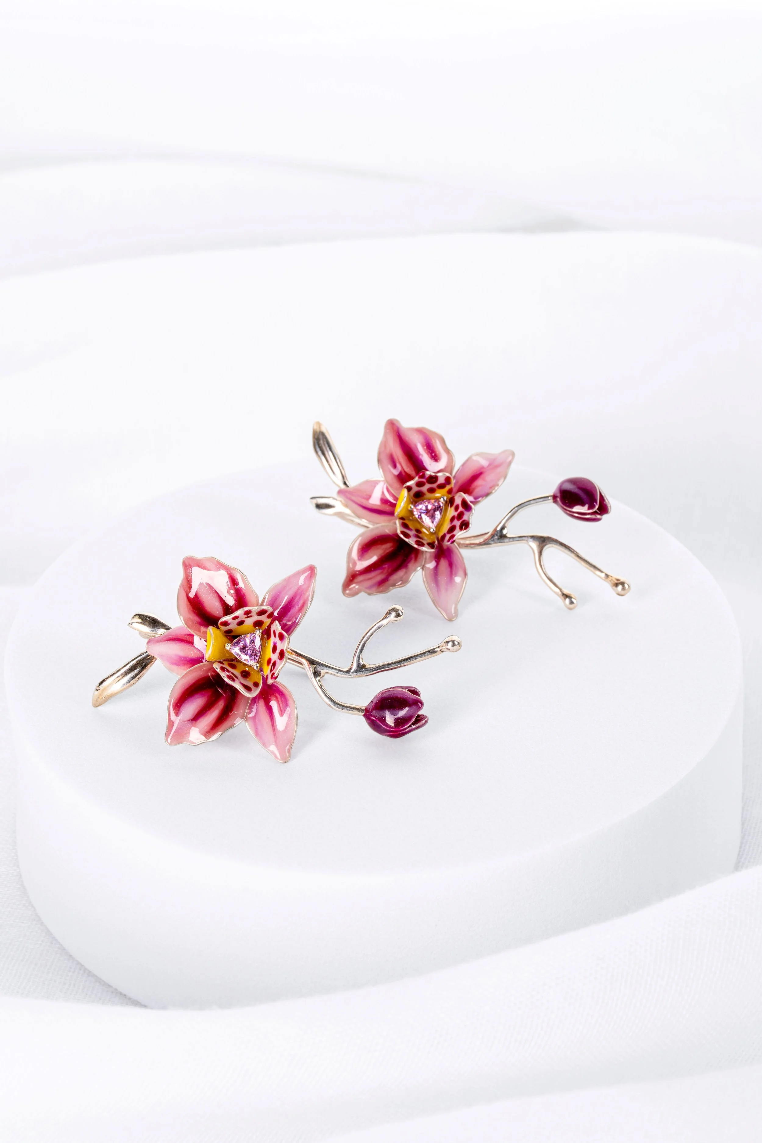 Pink flower-shaped brooches with gold accents and pink gemstones, arranged on a white display stand.