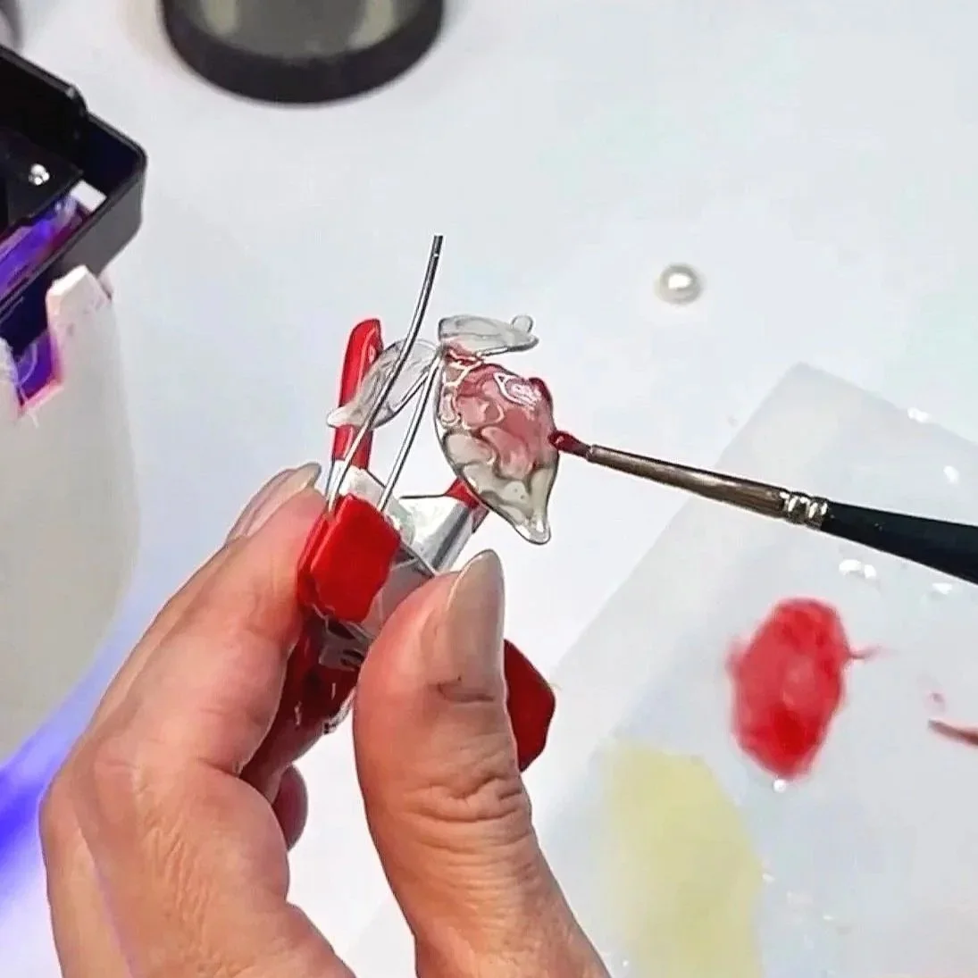 Person holding a delicate glass sculpture of a rose, using a small brush to add color or details, with art supplies and paper nearby.