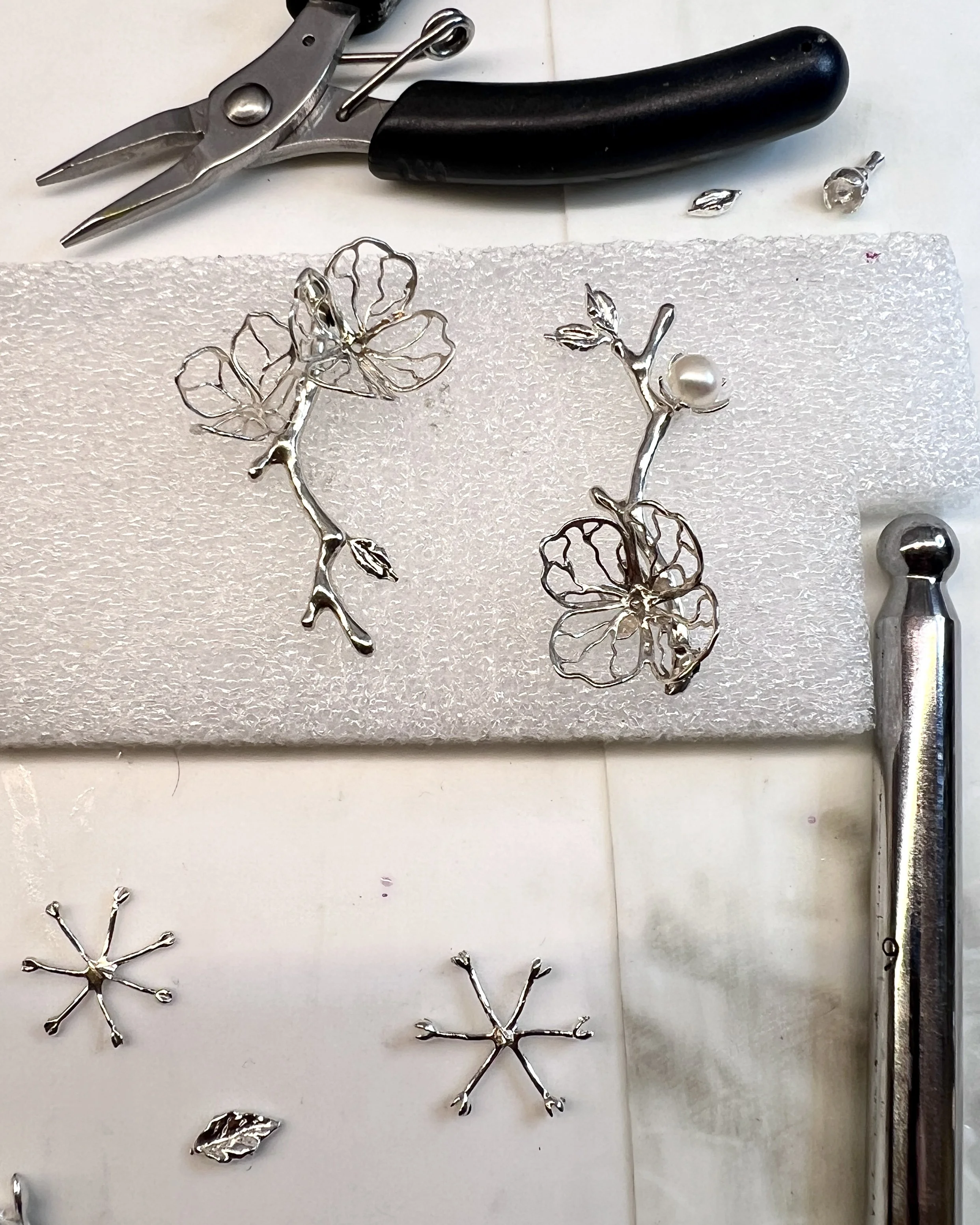 Silver floral jewelry pieces, including a butterfly, a branch with a pearl, and a flower, placed on white foam padding with jewelry tools nearby.
