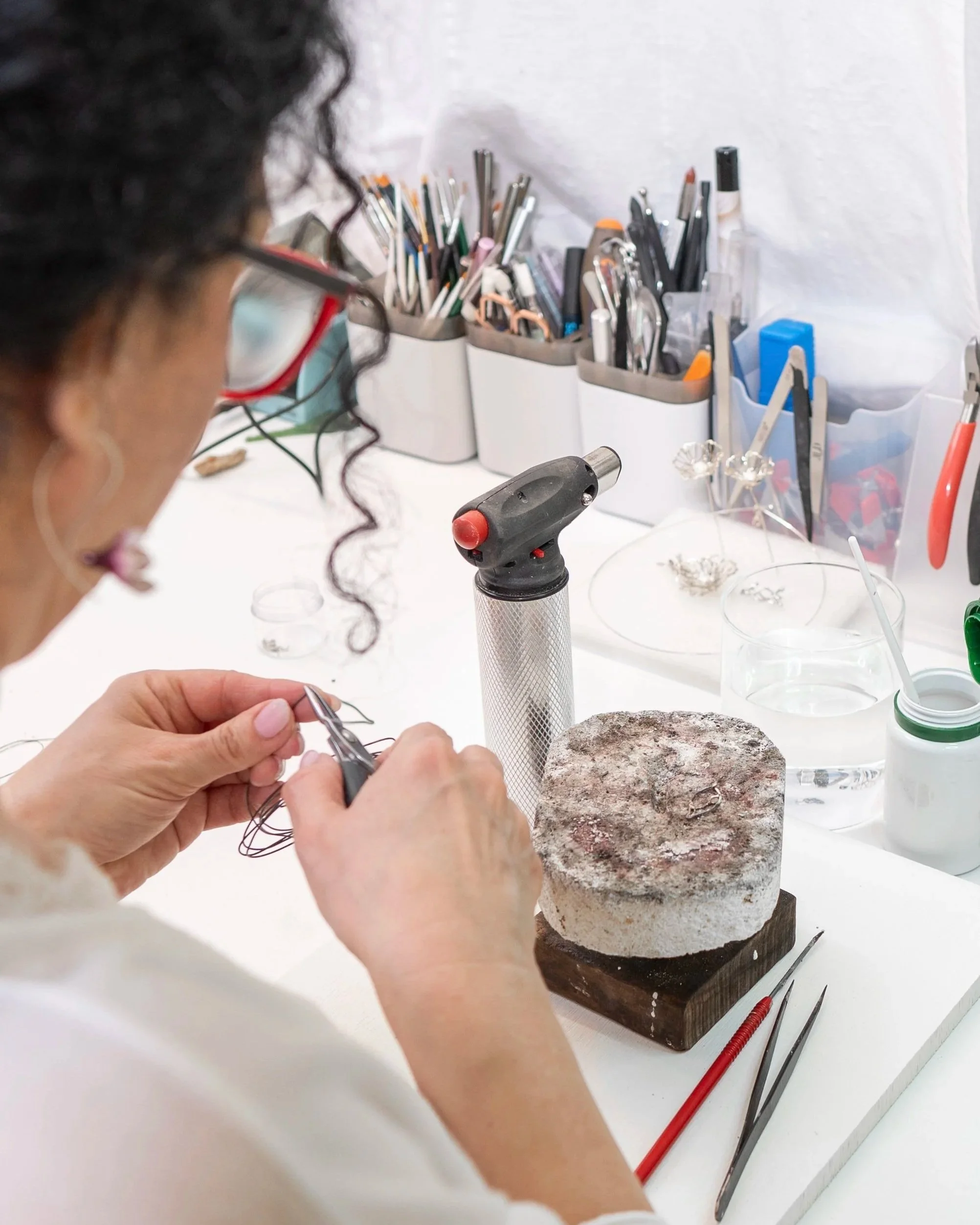A person working with jewelry tools at a workbench. Visible jewelry-making tools in organizers, a small torch, and a ring holder on a carved stone base.