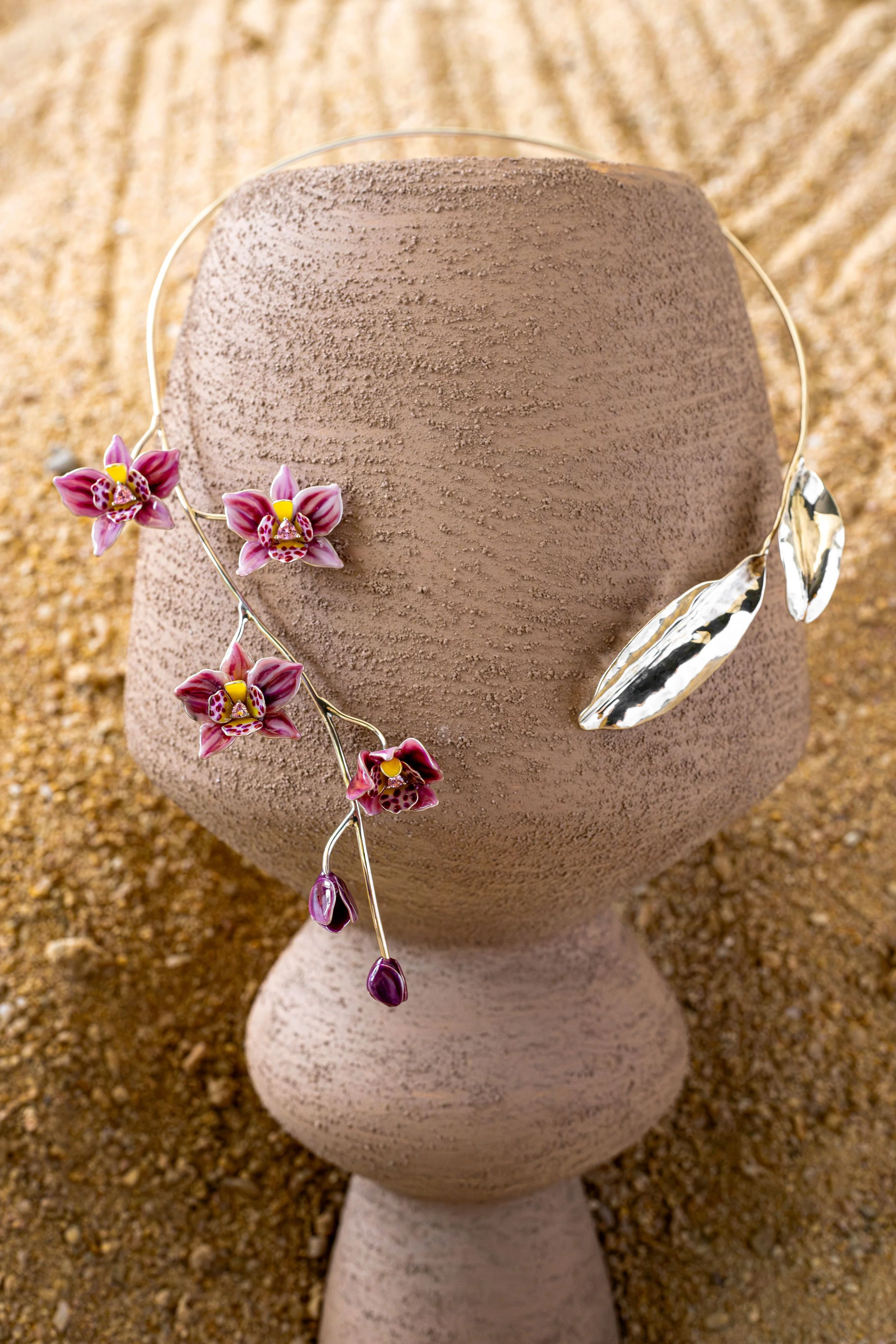 Decorative bust sculpture on a textured pedestal, adorned with a floral headpiece and metallic leaf accessories, set against a sandy background.