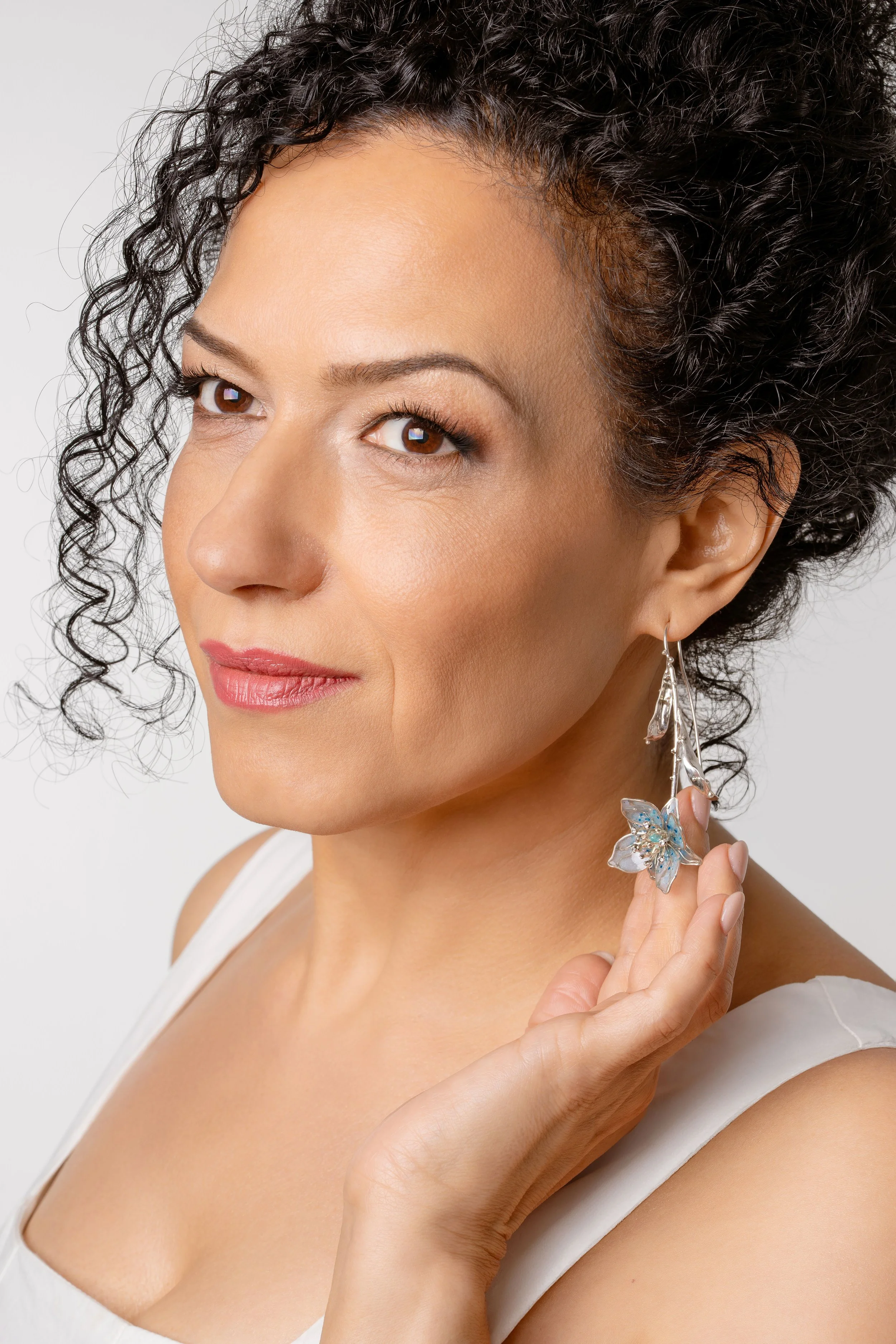 A woman with curly dark hair wearing a white top, holding a butterfly-shaped earring with her right hand, looking slightly to her left with a neutral expression.