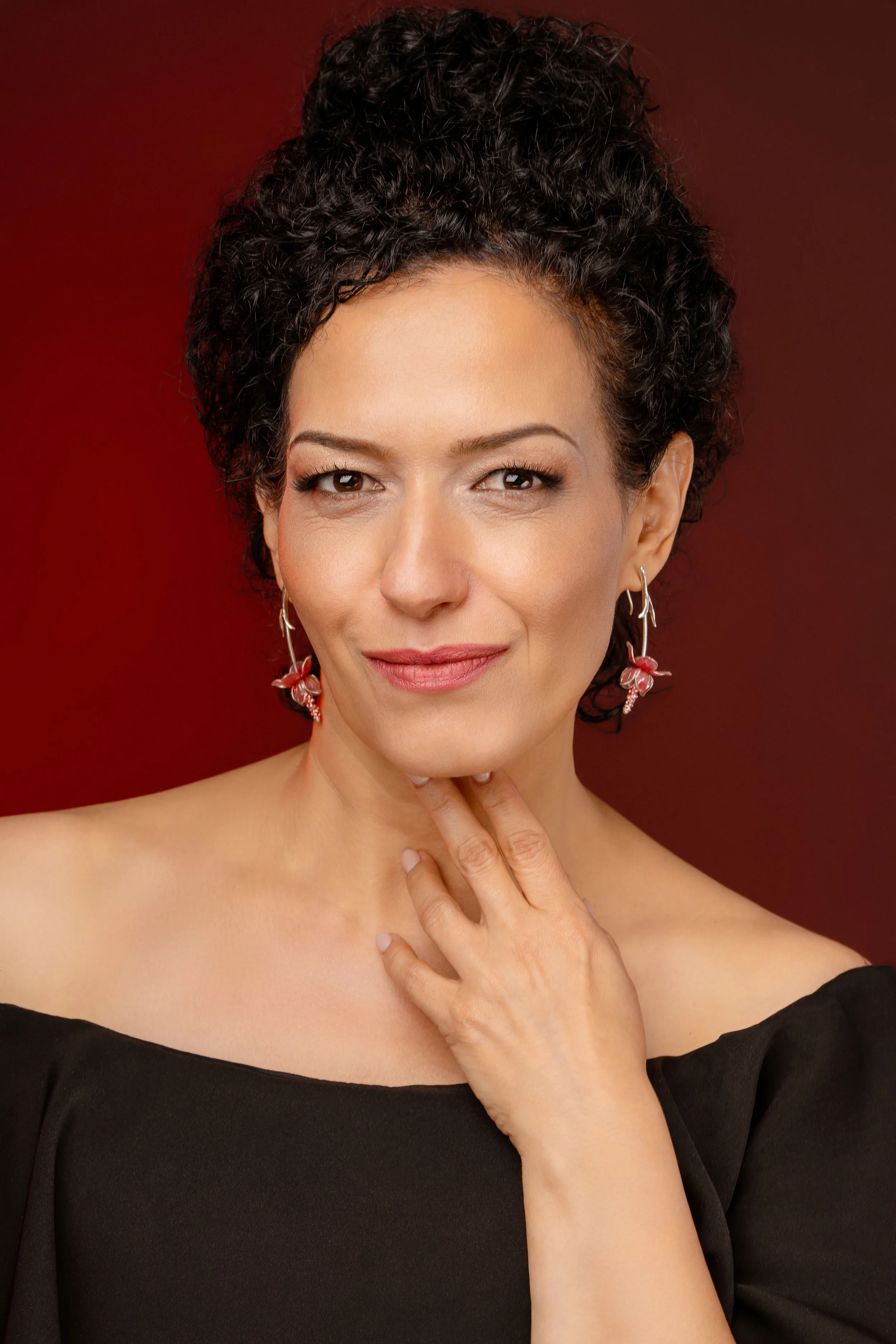 A woman with dark curly hair, wearing earrings and an off-shoulder black top, is smiling softly with her hand touching her neck against a red background.