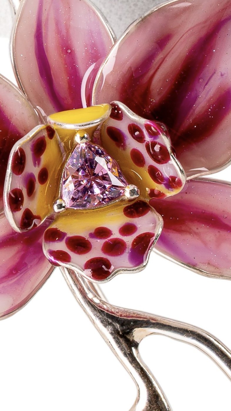 A decorative brooch in the shape of a flower with pink petals, a yellow and pink-dotted center, and a pink gemstone in the middle, set in silver.